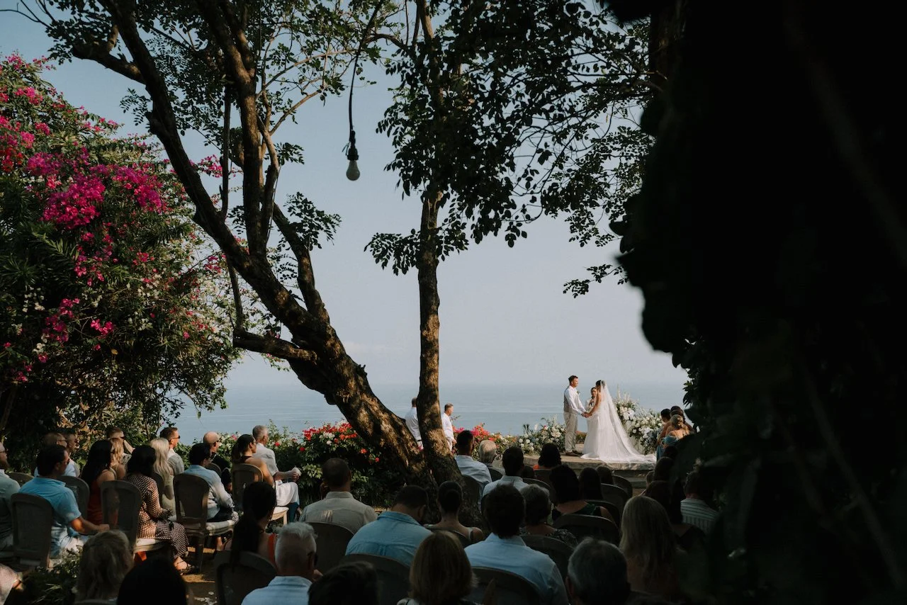 Clifftop wedding ceremony at Wonderland seen through trees and pink bougainvillea, with the ocean behind