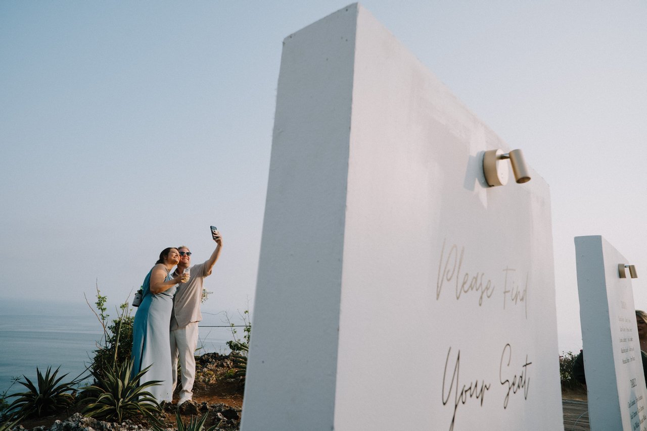 Wedding guests taking a selfie beside a white “Please Find Your Seat” sign above the ocean