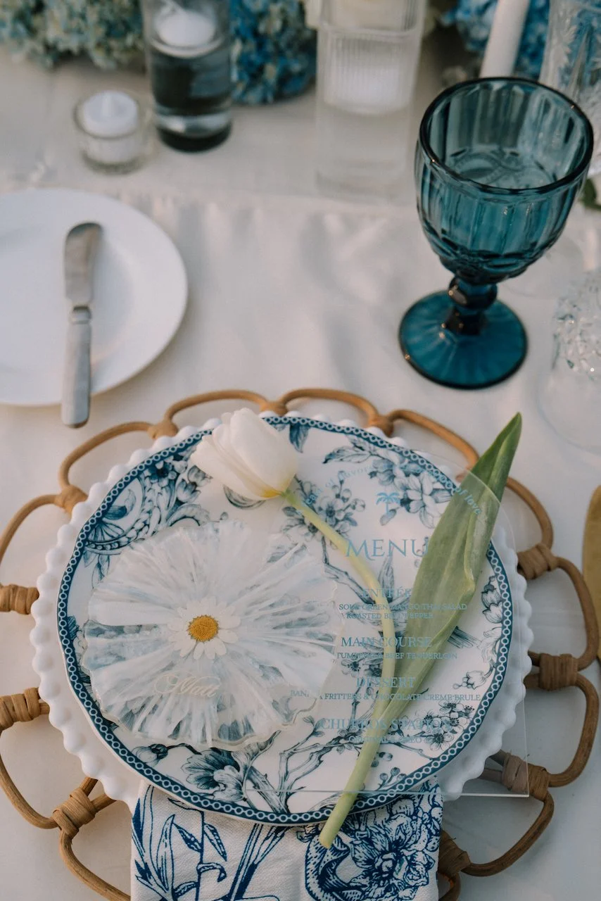 Blue and white floral plate on a scalloped white plate and rattan charger, with a white tulip, acrylic menu and daisy place card for Matt