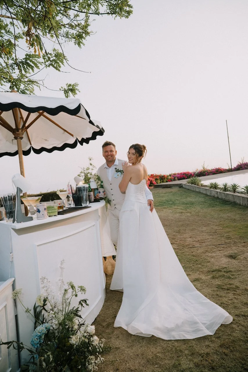 Matt and Kelly leaning on a white bar under a black-trimmed umbrella with cocktails, her dress train spread on the grass