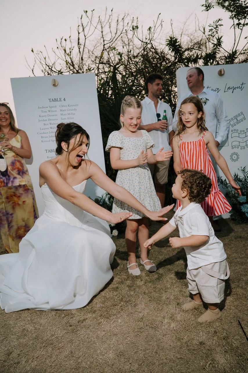 Kelly crouching with open arms to greet a toddler as two young girls and guests look on beside the seating plan