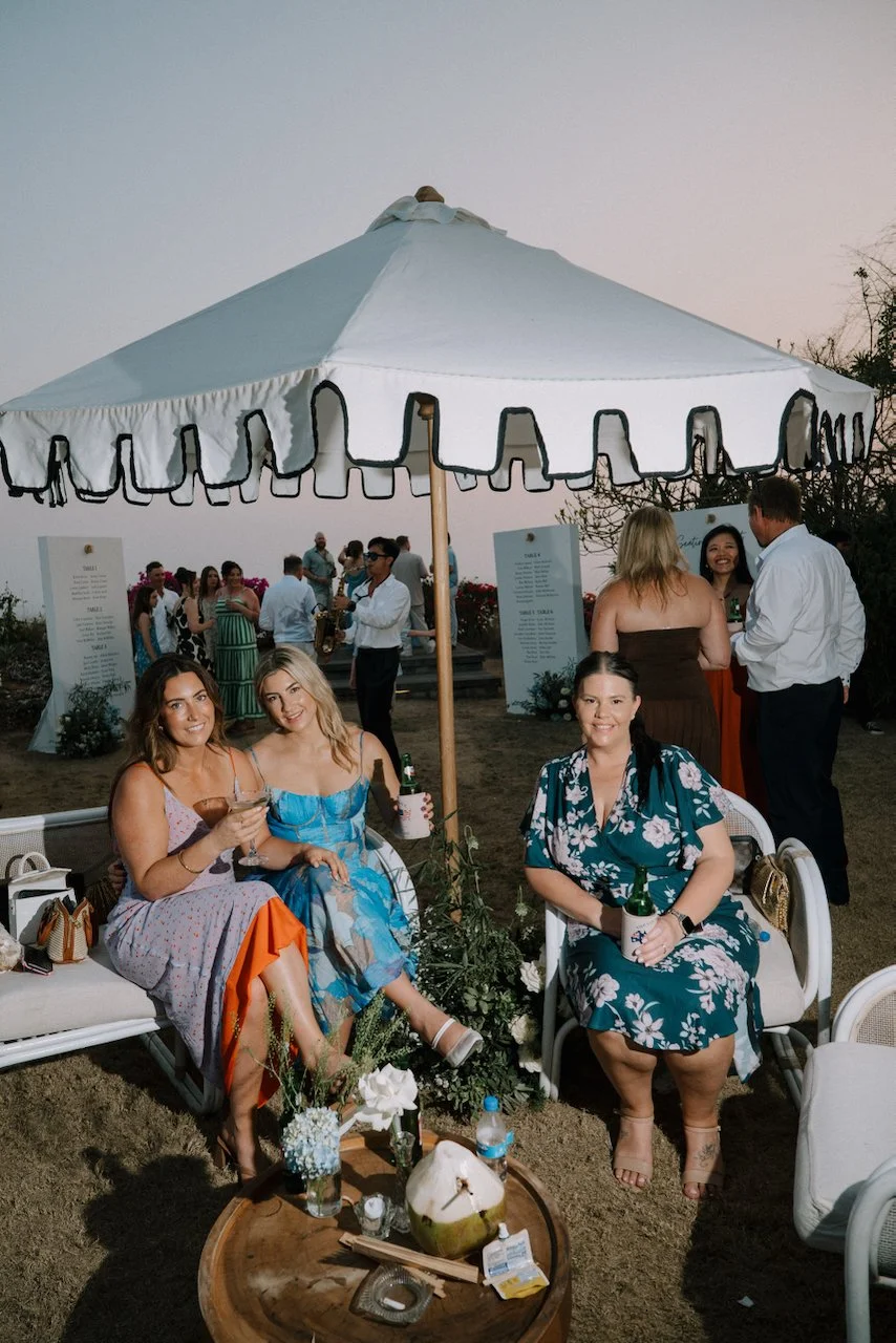 Three guests seated under a scalloped umbrella with beers and cocktails, with a saxophonist playing behind them