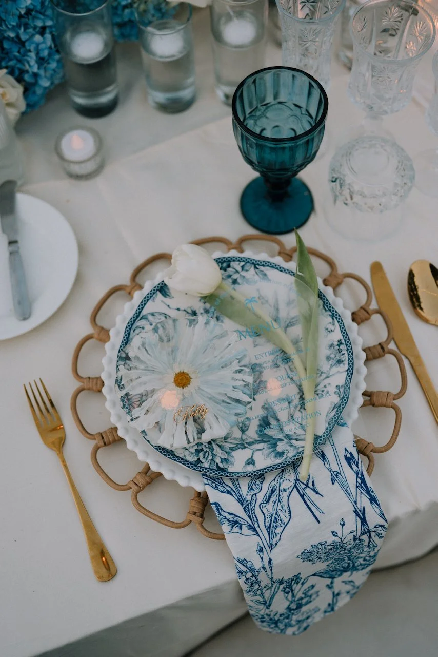 Place setting with gold cutlery, a toile napkin, a white tulip and a daisy name card for Kelly beside a teal goblet