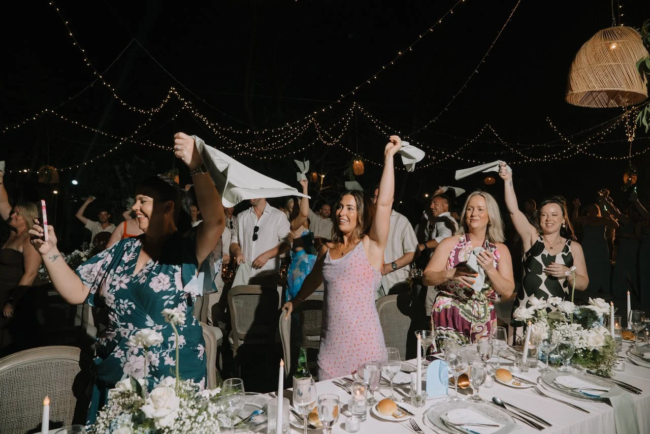 Wedding guests standing at the reception tables and waving napkins under festoon lights and rattan lanterns