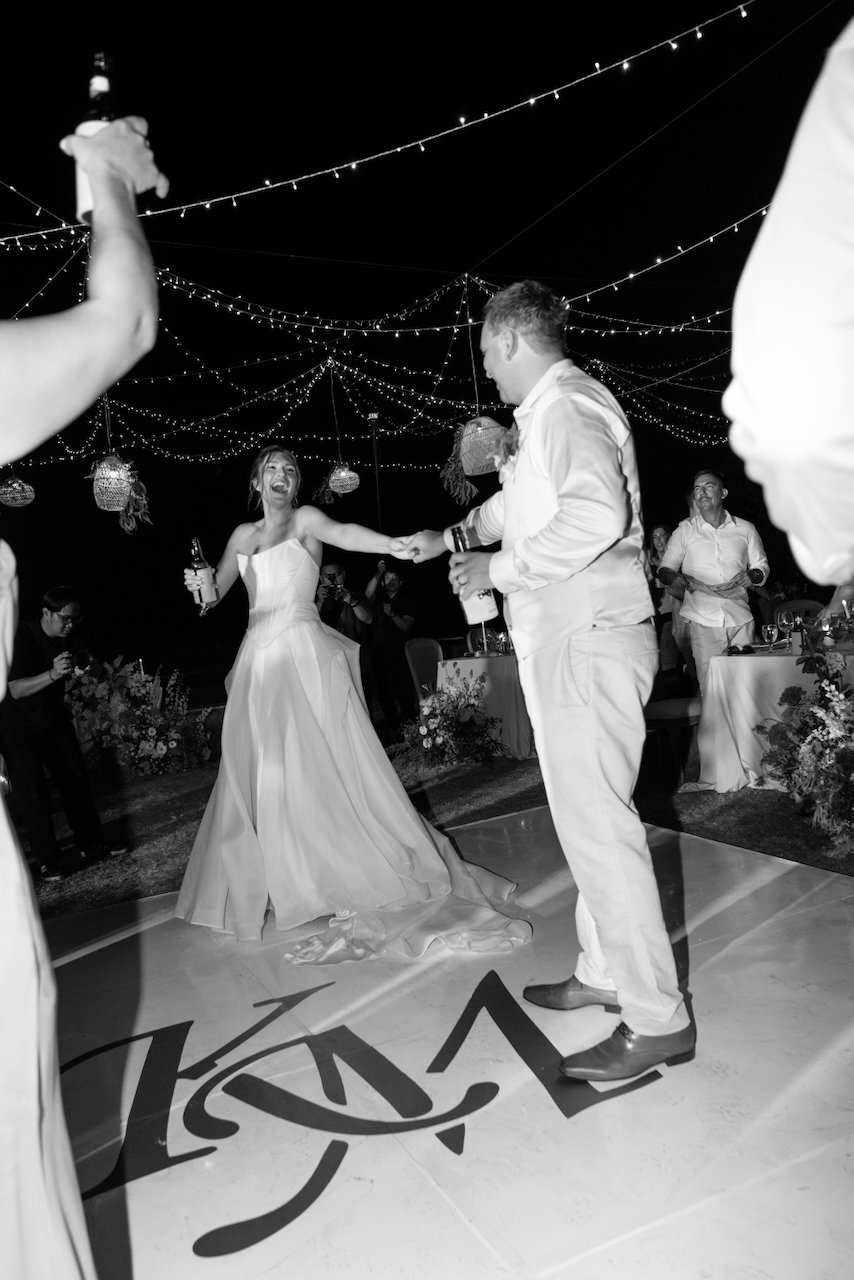 Black and white photo of Kelly laughing as Matt takes her hand on the monogrammed dance floor, guests and photographers watching