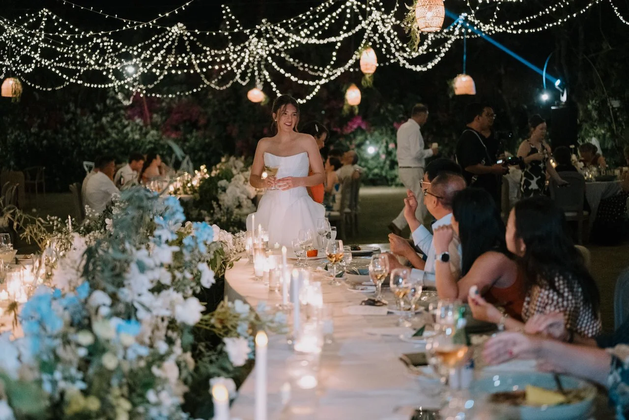 Kelly smiling at the candlelit bridal table, holding a glass, with blue and white flowers and guests clapping