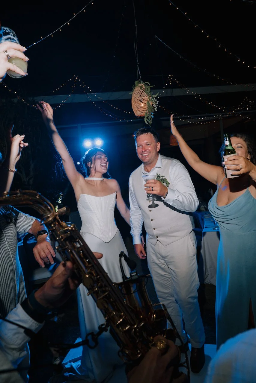 Kelly, Matt and a bridesmaid dancing and cheering with drinks as a saxophone plays in the foreground