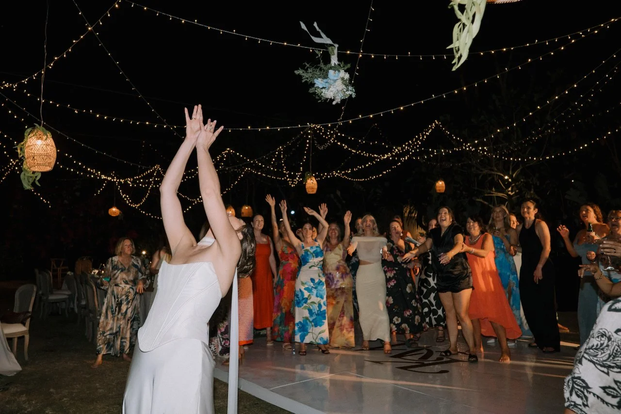 Kelly throwing her bouquet to guests with their hands raised under the lights and hanging rattan lanterns