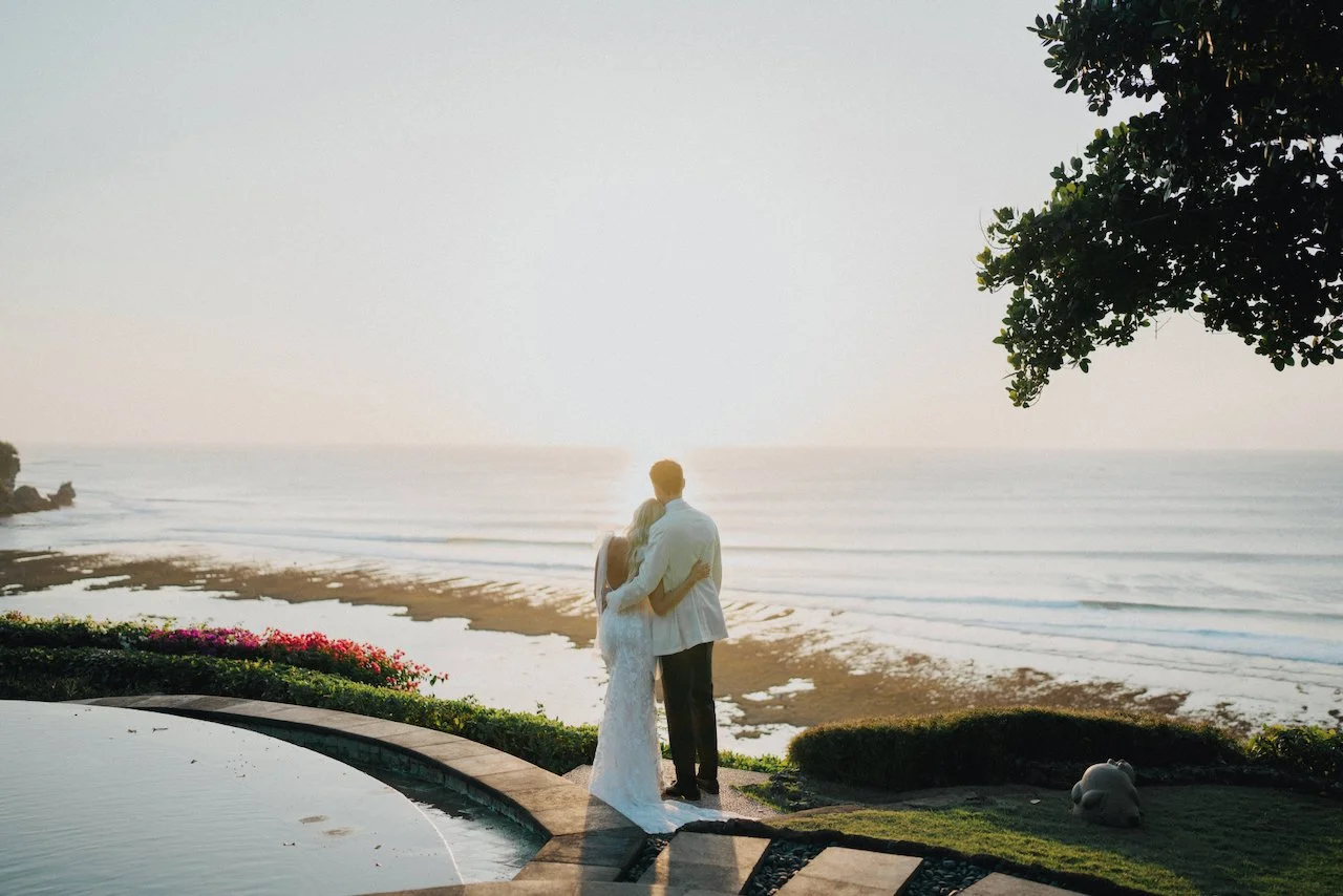 Bride and groom embracing while overlooking the ocean at sunset during a luxury Bali destination wedding, photographed at a clifftop villa with coastal views and tropical gardens.