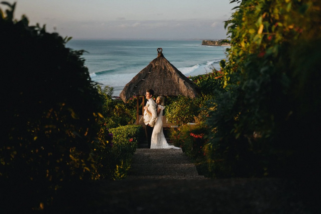 Bride and groom standing together at a cliffside Bali wedding venue overlooking the ocean, surrounded by tropical gardens.