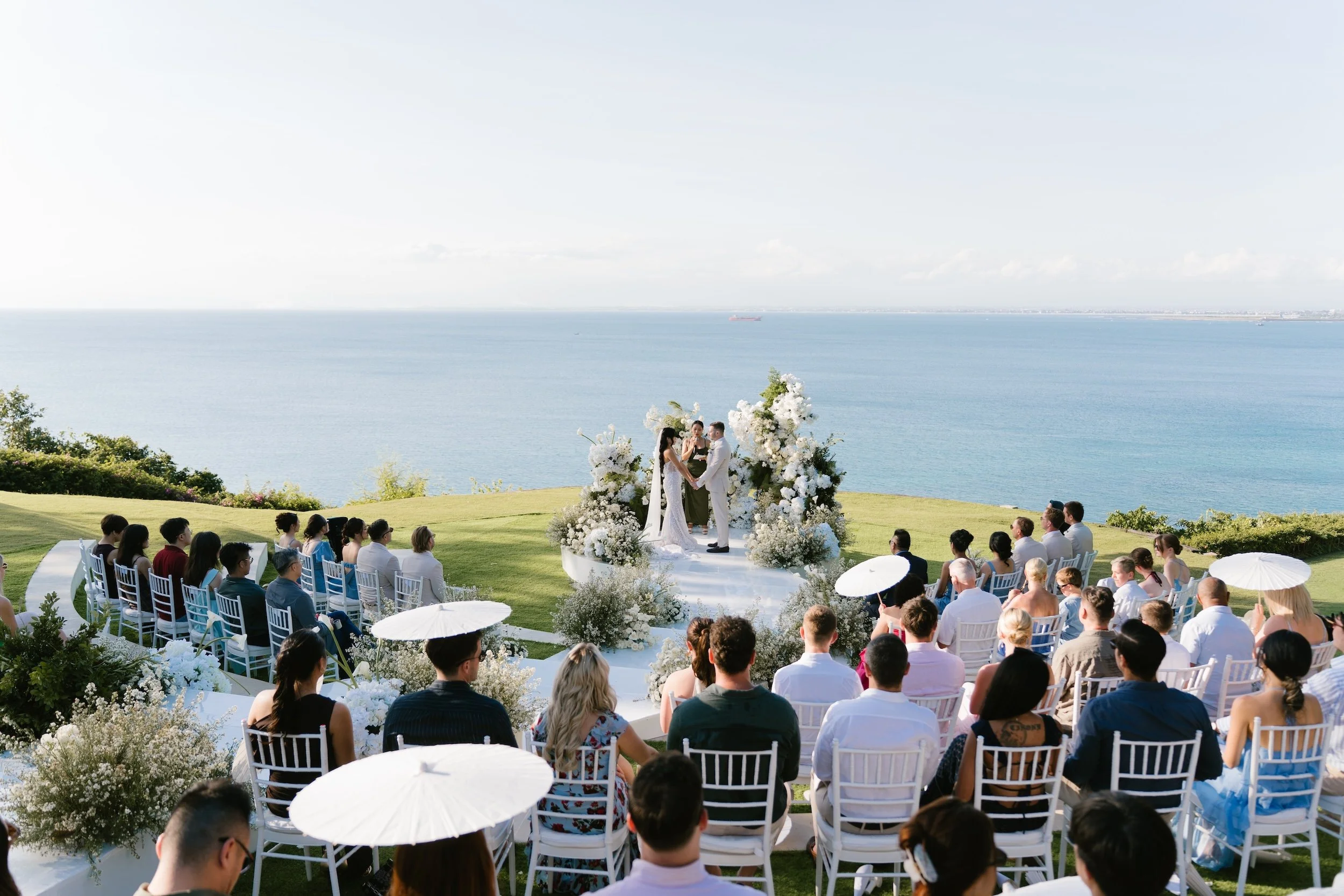 Outdoor cliffside wedding ceremony at AYANA Resort in Bali with a white floral altar overlooking the ocean, planned by Silver Lace Weddings