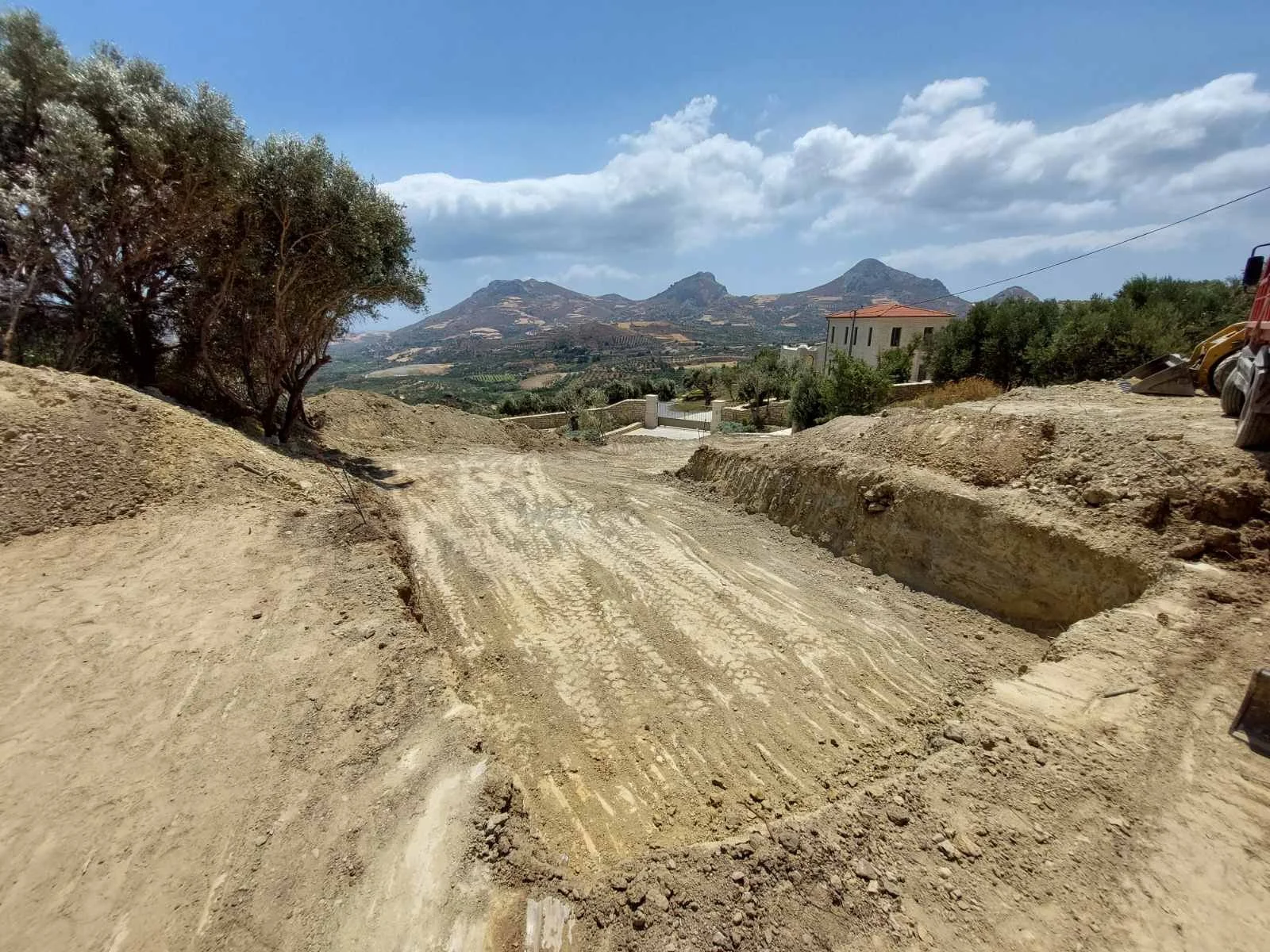 A dirt road under construction with tire tracks, on a hill with a tree on the left and a house on the right, mountains and partly cloudy sky in the background.