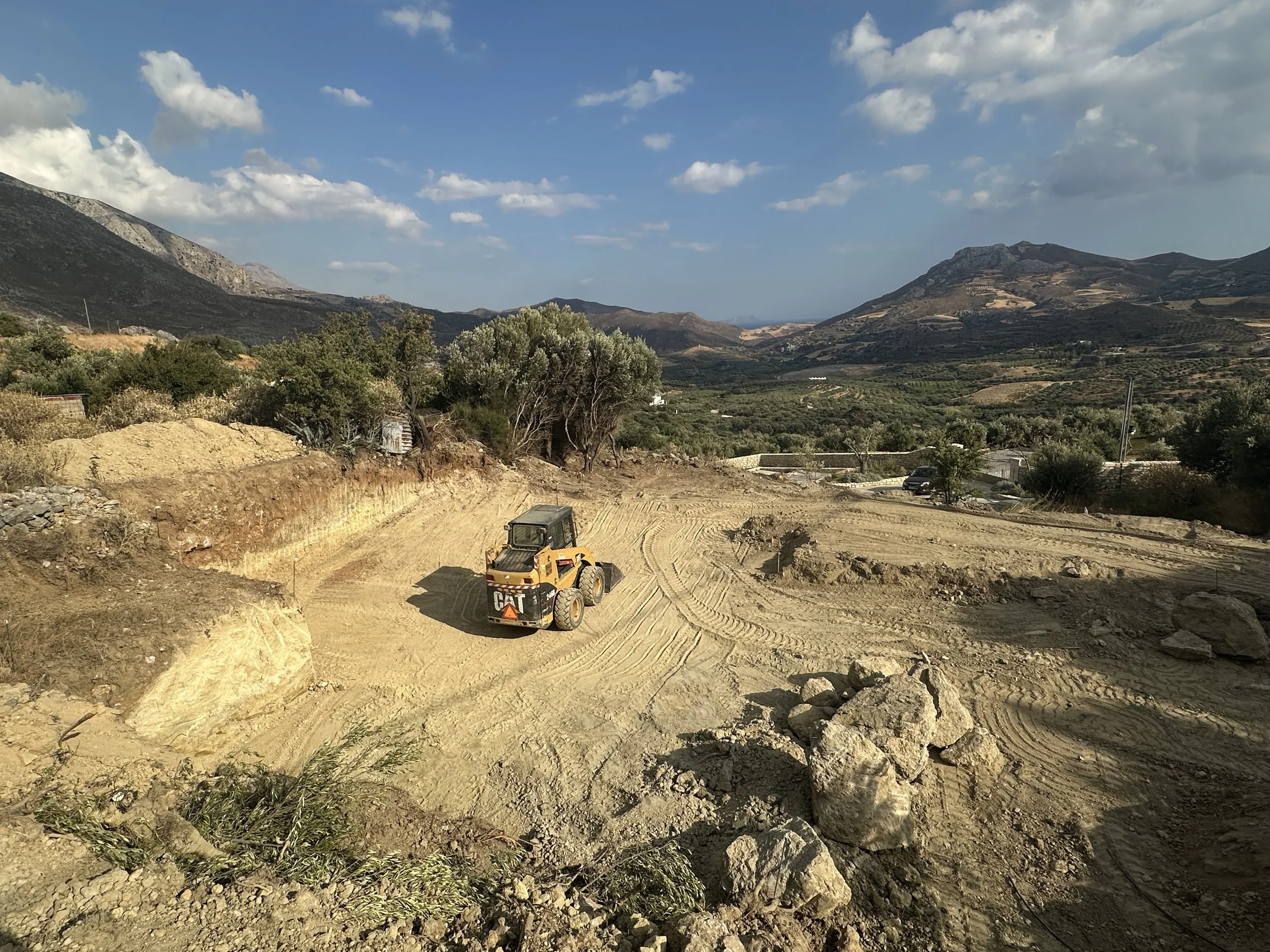 Construction site with a yellow CAT bulldozer on dirt, surrounded by rocks and sparse vegetation, with hills and mountains in the background under a partly cloudy sky.