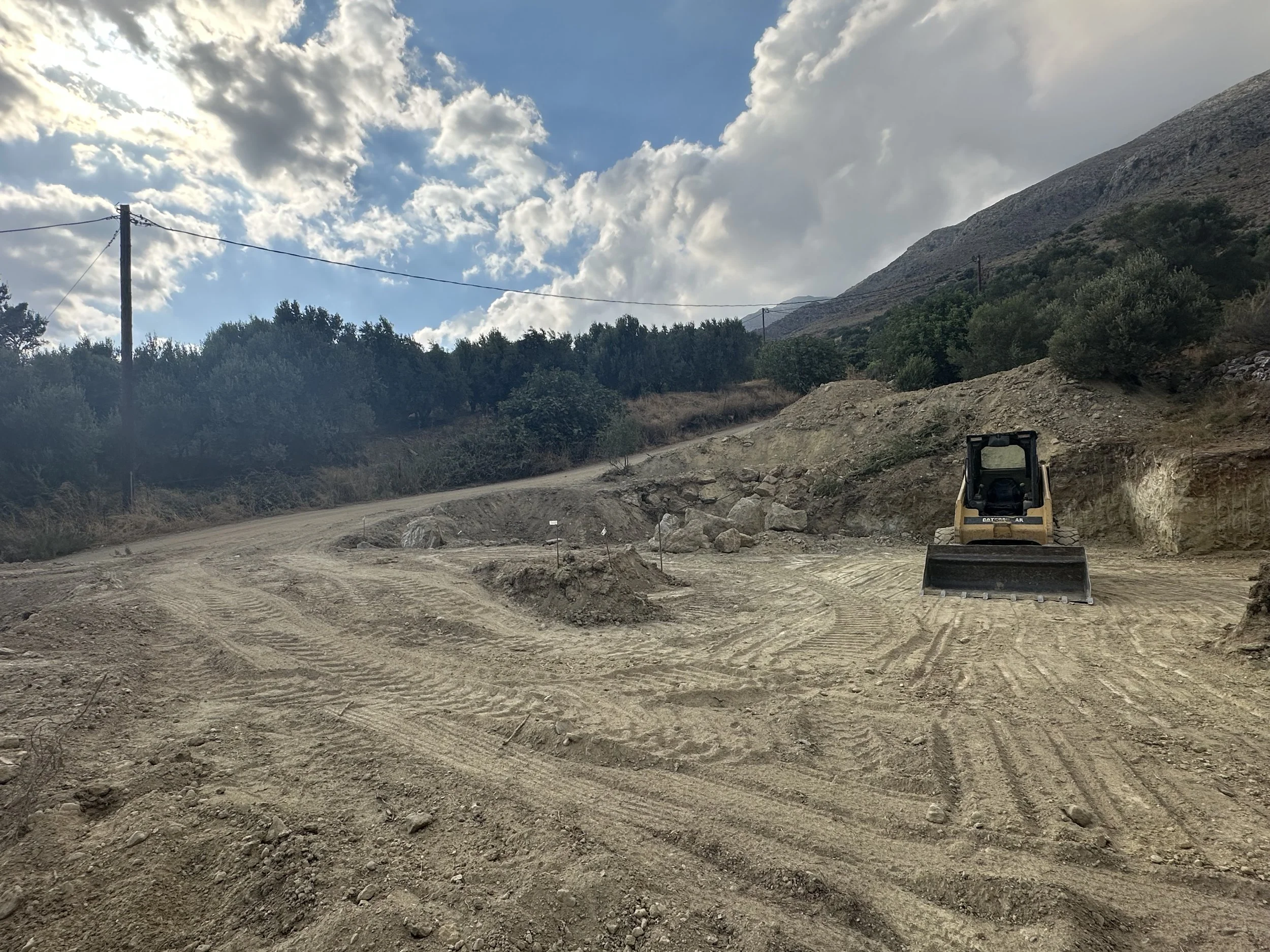 A construction site with a bulldozer on a dirt road, surrounded by hills and trees, under a partly cloudy sky.