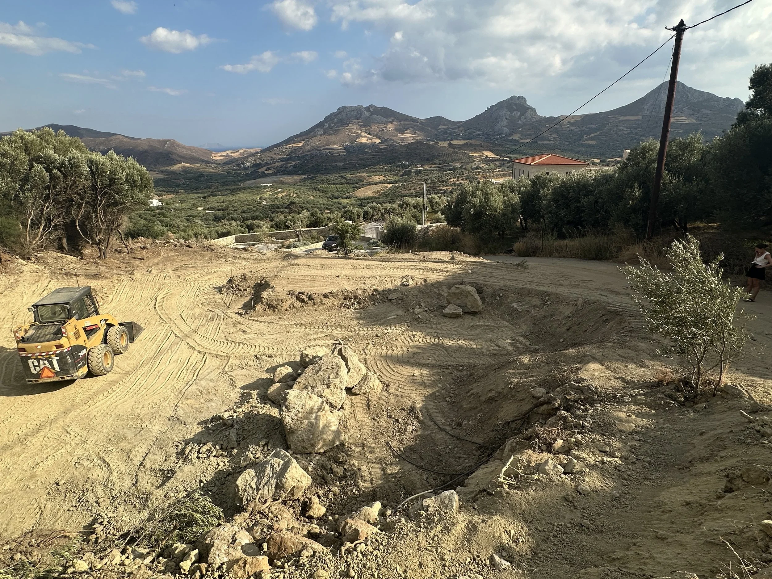 Rough dirt road with tire tracks, large rocks, construction vehicle, and a person standing near the edge, with mountains and partly cloudy sky in the background.