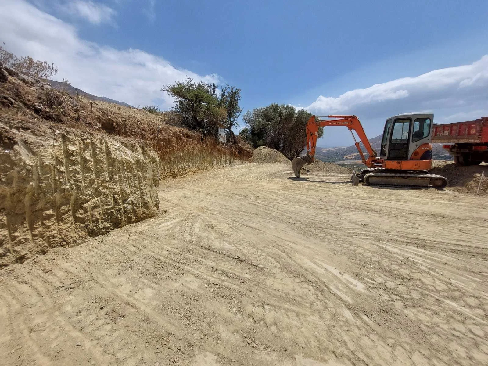 A construction site with an orange mini excavator digging on a dirt road, with a truck parked nearby. The landscape includes a rocky hillside with sparse trees under a partly cloudy sky.