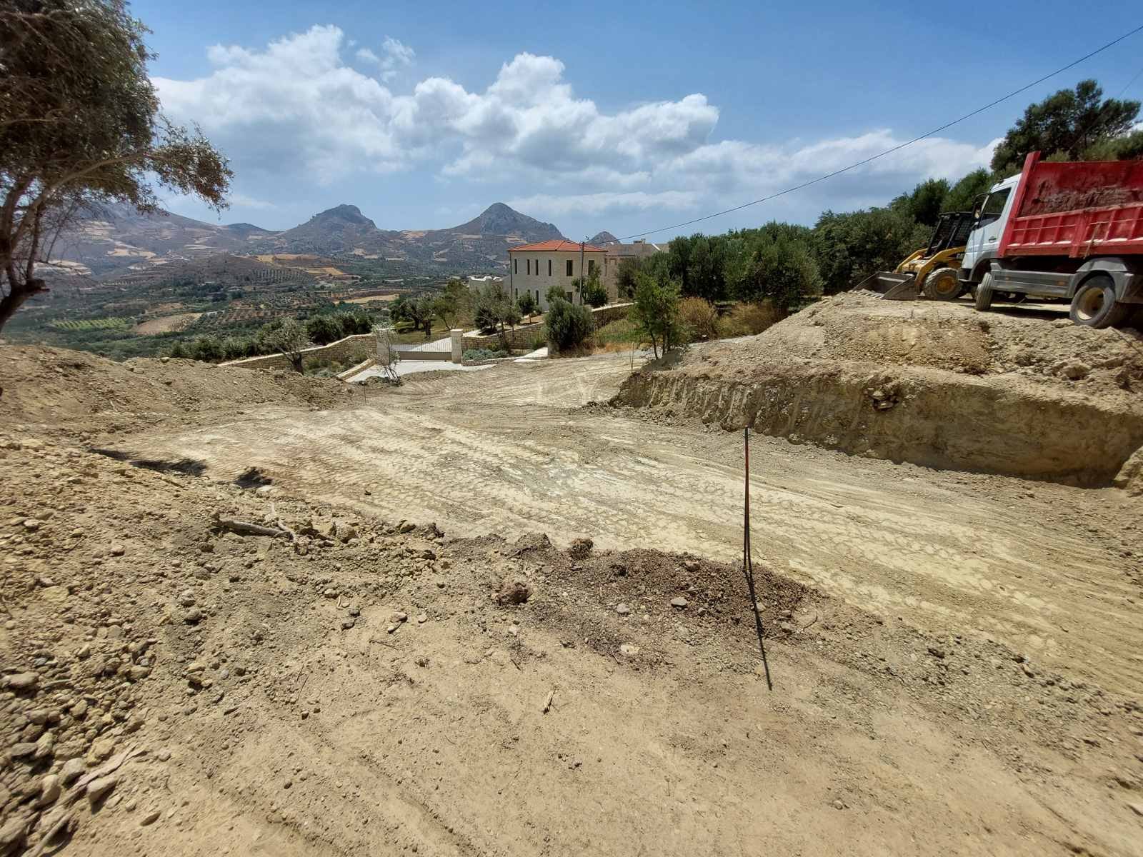 Construction site on a dirt road with construction vehicles, trees, a house, and mountains in the background under a partly cloudy sky.