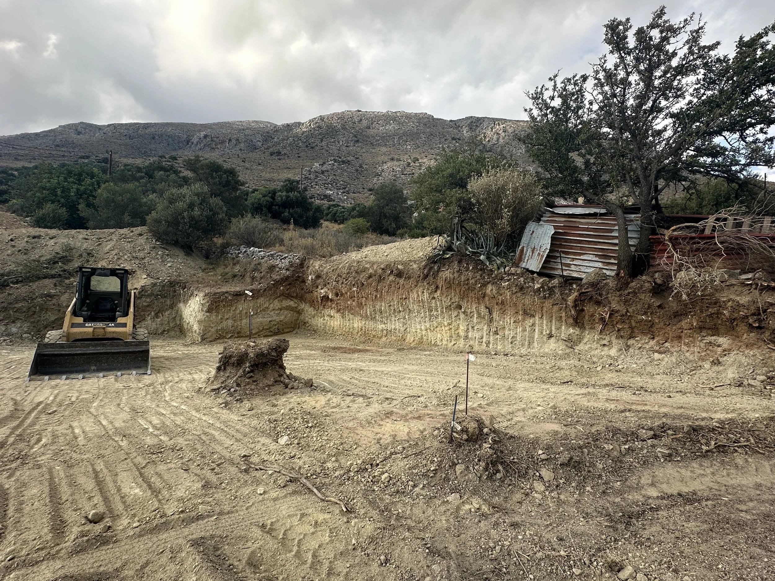 Construction site with a small bulldozer, cleared dirt, and a hillside with sparse vegetation and a rusted metal shed in the background.