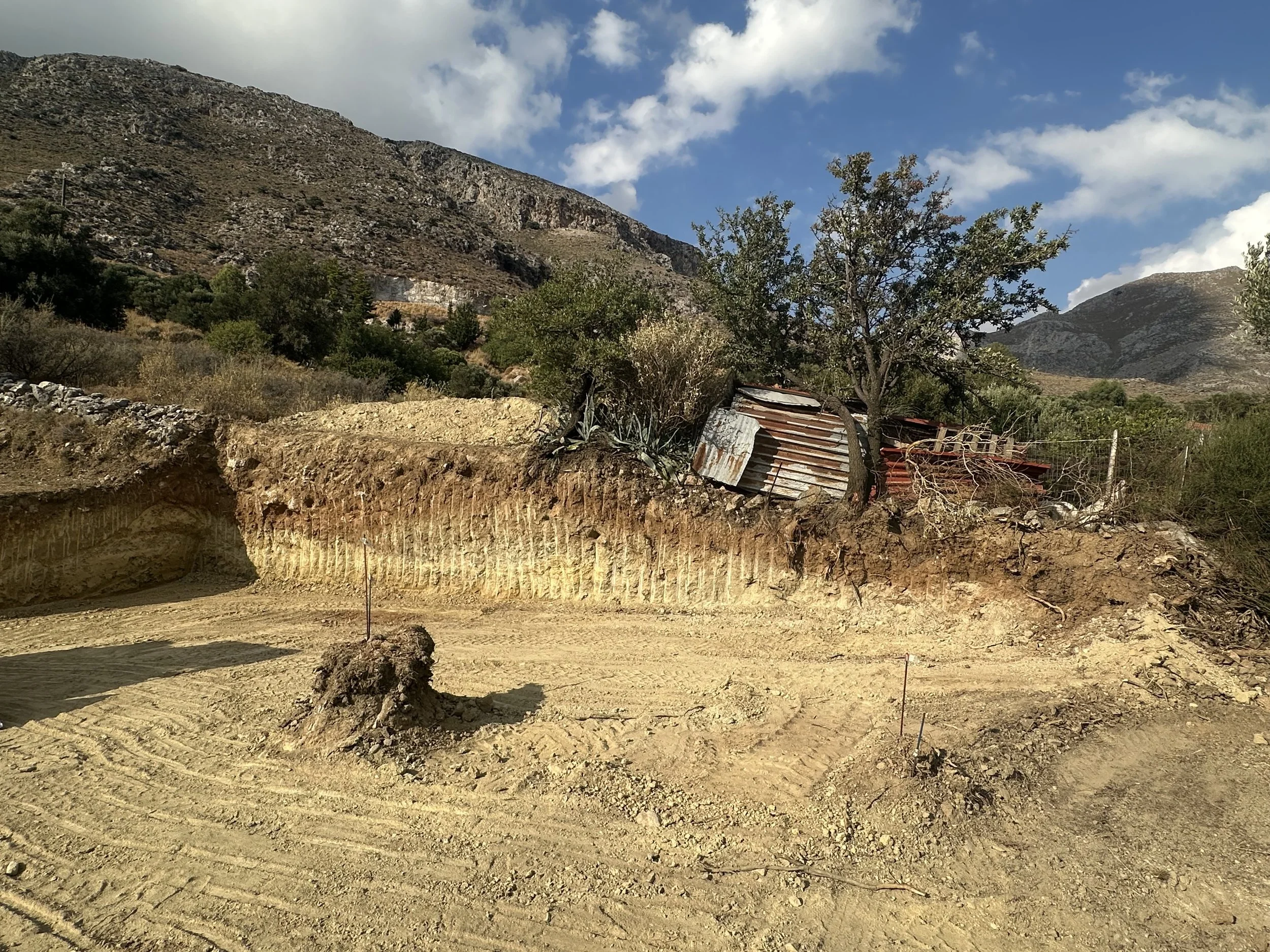 A dirt construction site with a small tree and a rusty shed in a hilly, semi-arid landscape with mountains and a partly cloudy sky in the background.
