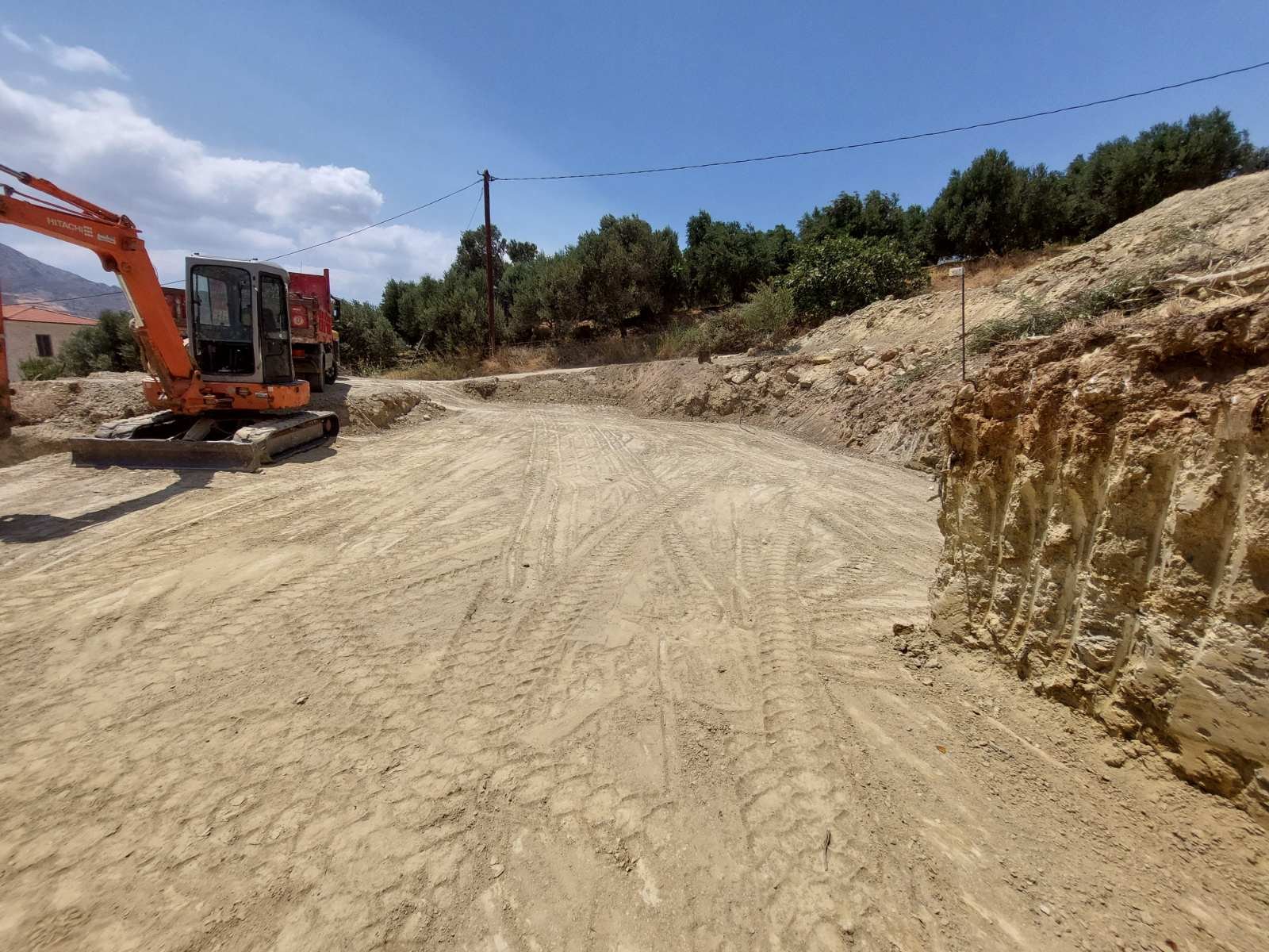 Construction site on a dirt road with an orange excavator parked, surrounded by trees, utility poles, and a hill under a partly cloudy sky.