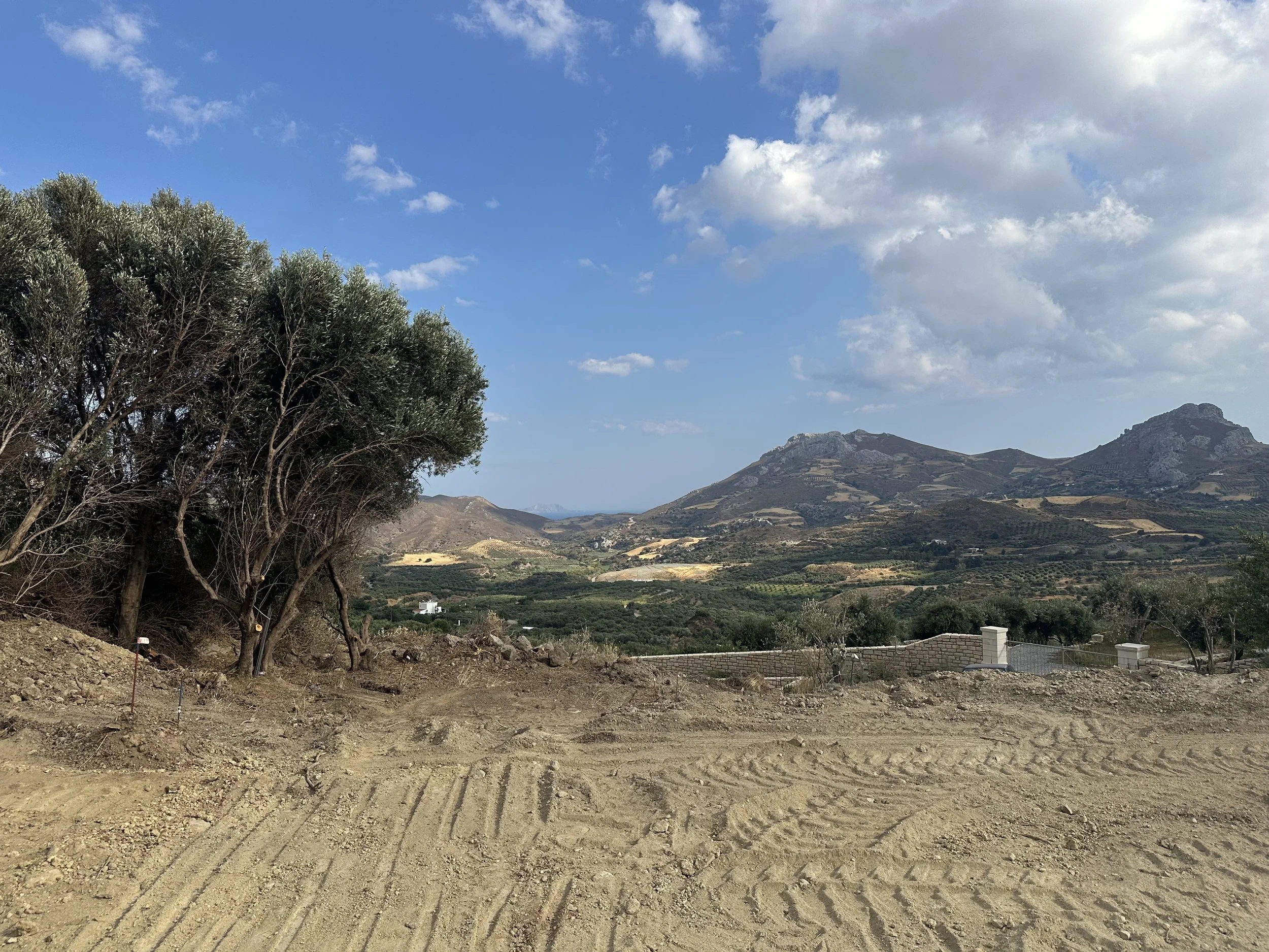 A dirt road in a mountainous landscape with trees on the left and a view of hills and mountains under a partly cloudy sky.