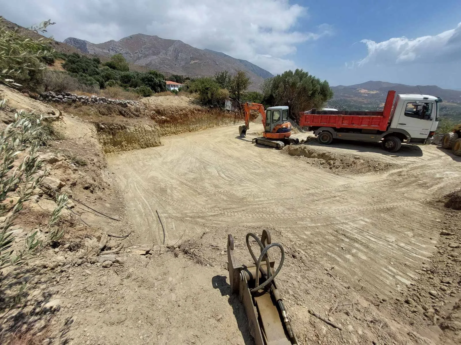 Construction site on a dirt road with a small excavator and a dump truck, mountainous landscape in the background, cloudy blue sky.