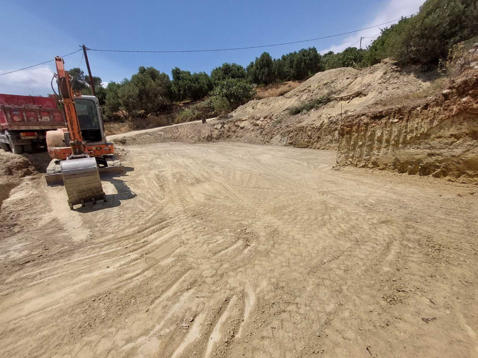 Construction site with an orange mini excavator next to a red dump truck on a dirt road, with a hillside and trees in the background under a blue sky.
