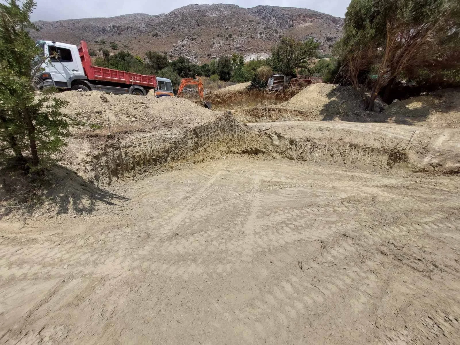 A dirt construction site with two trucks, an orange excavator, and a mound of dirt. Hills and sparse trees are in the background.