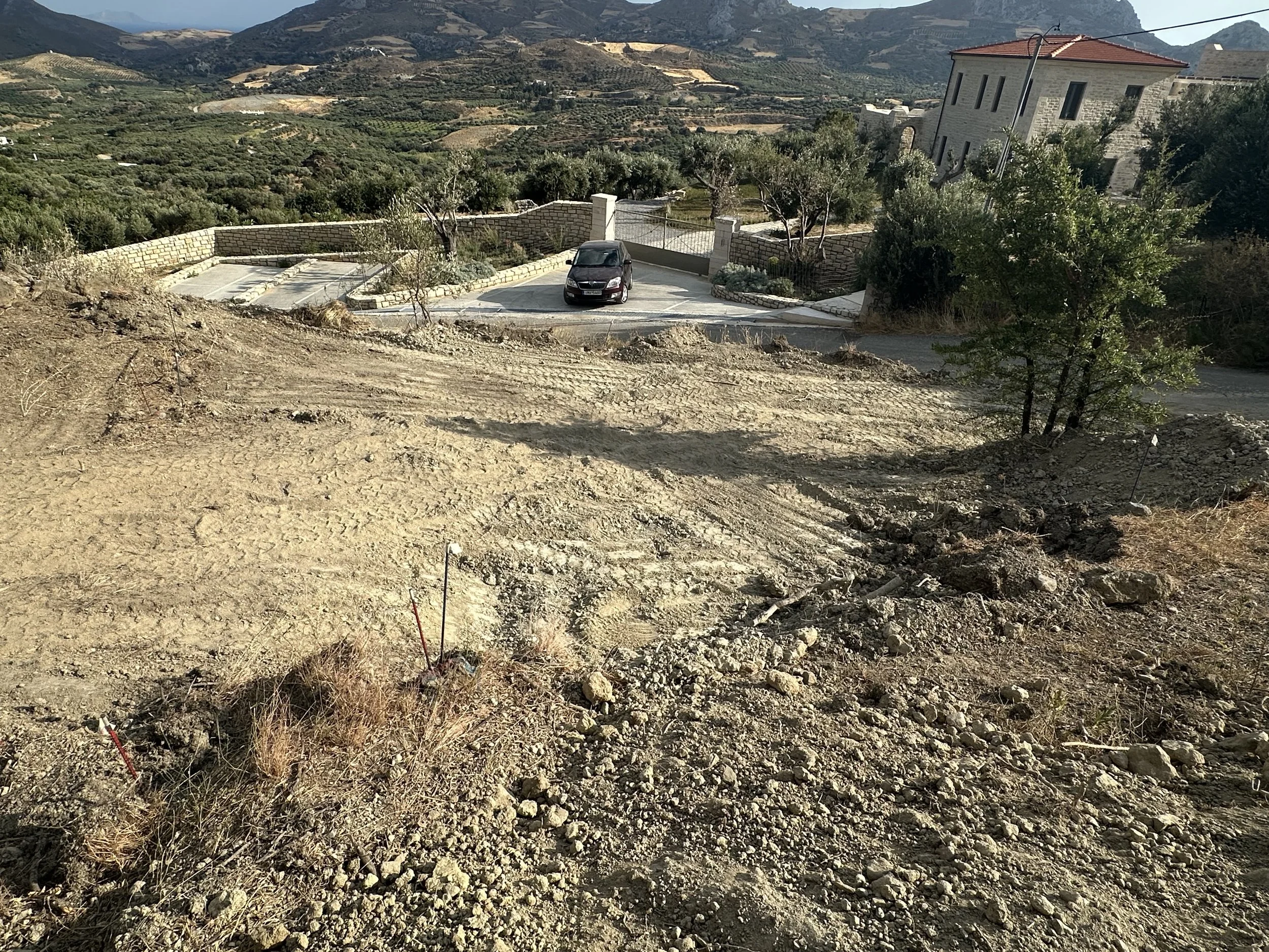 Une zone en construction ou en terrassement sur un terrain en pente, avec des marques de travaux, près d'une maison en pierre et d'une voiture, avec un paysage montagneux en arrière-plan.