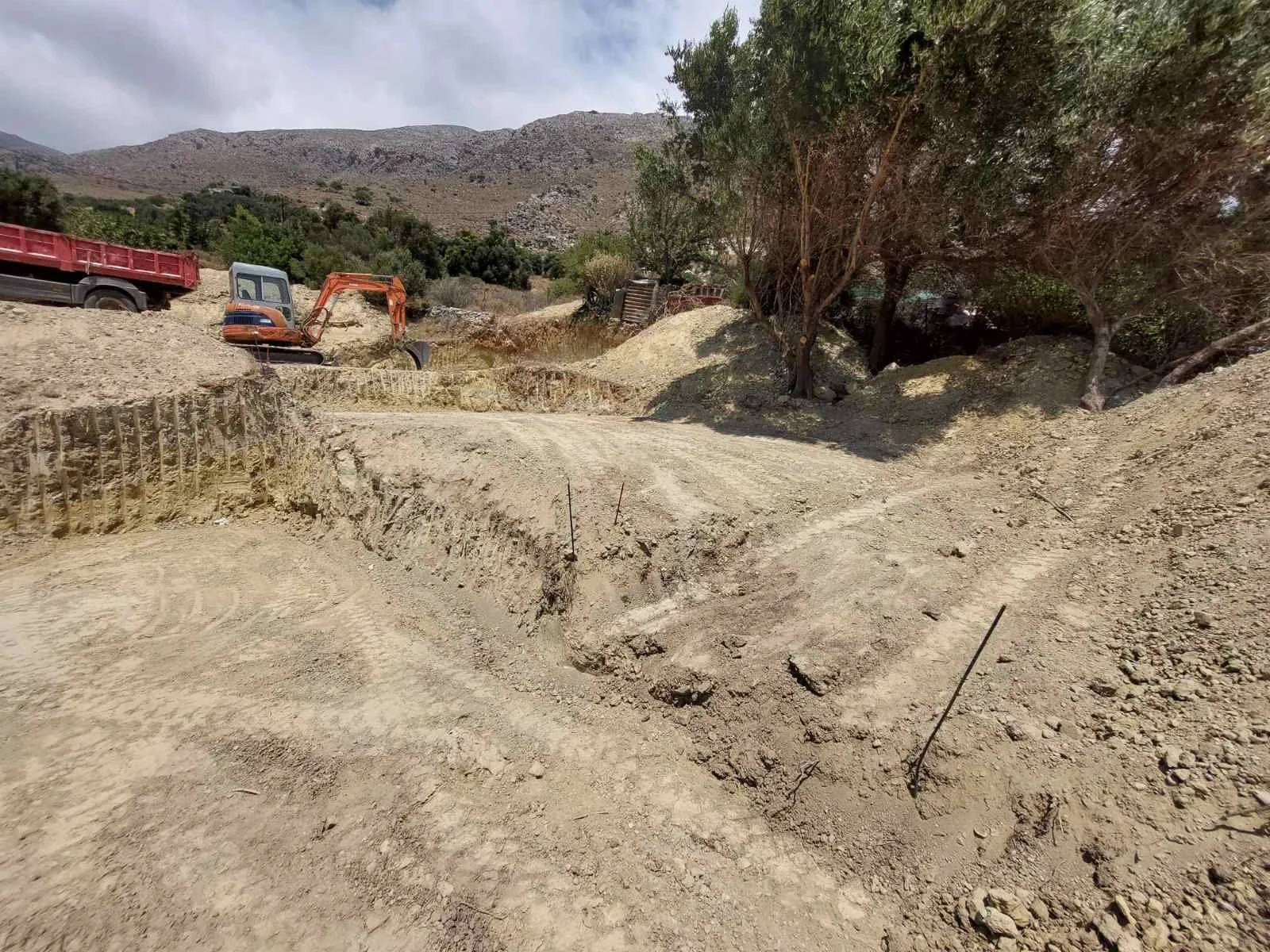 A construction site with dirt and a small excavator, trees, and mountains in the background.