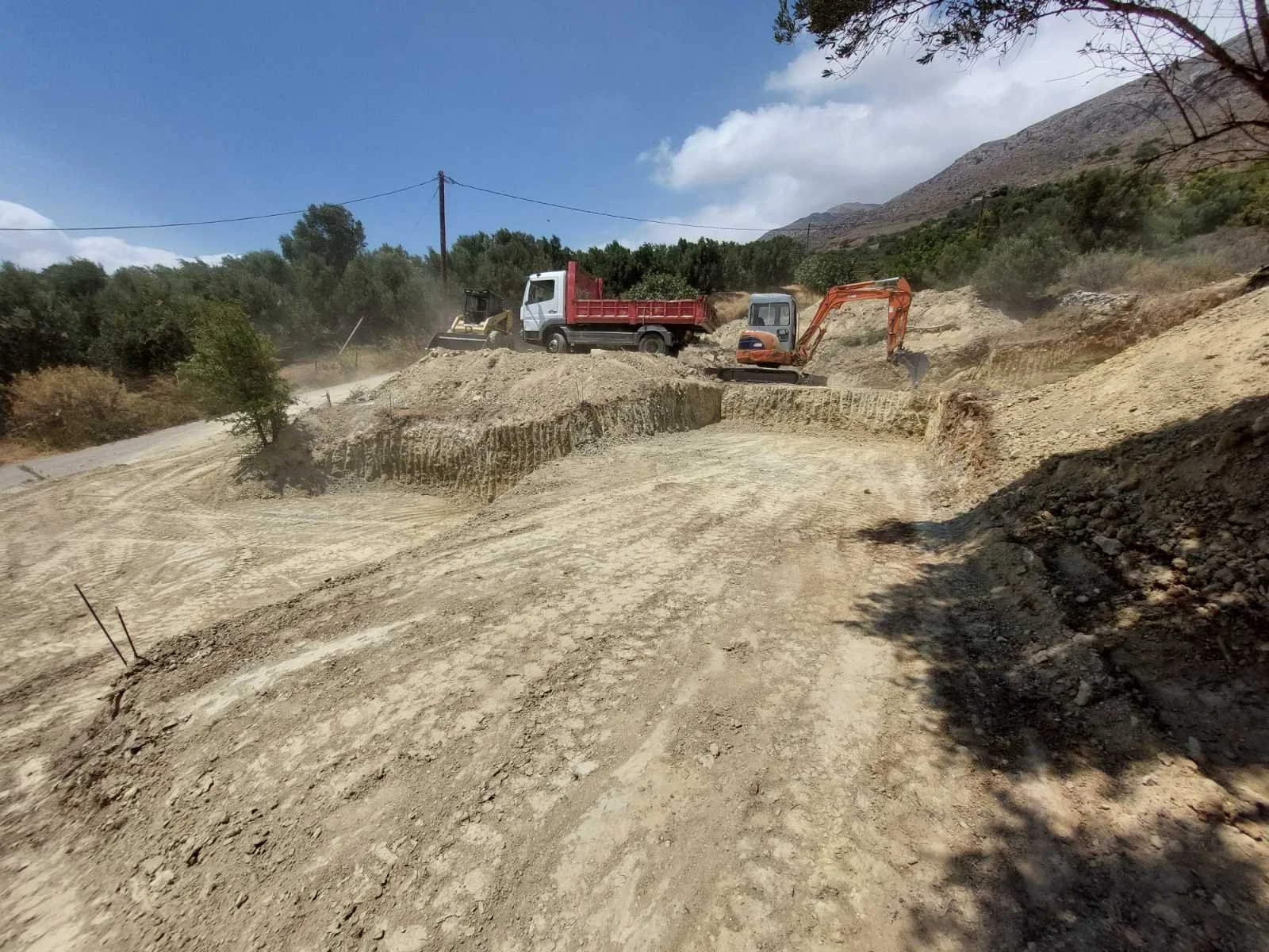 Construction site on a dirt road with dump truck, excavator, and other machinery working, mountains and trees in the background, partly cloudy sky.