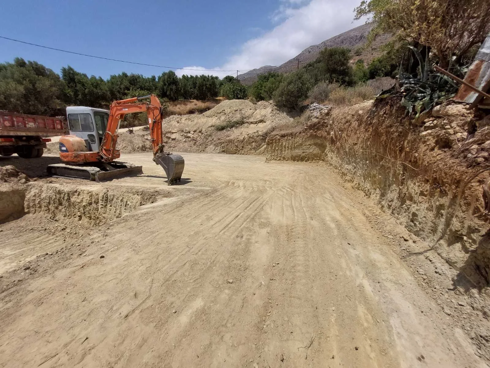Construction site with an orange mini excavator on a dirt road, surrounded by rocky terrain and hills in the background.