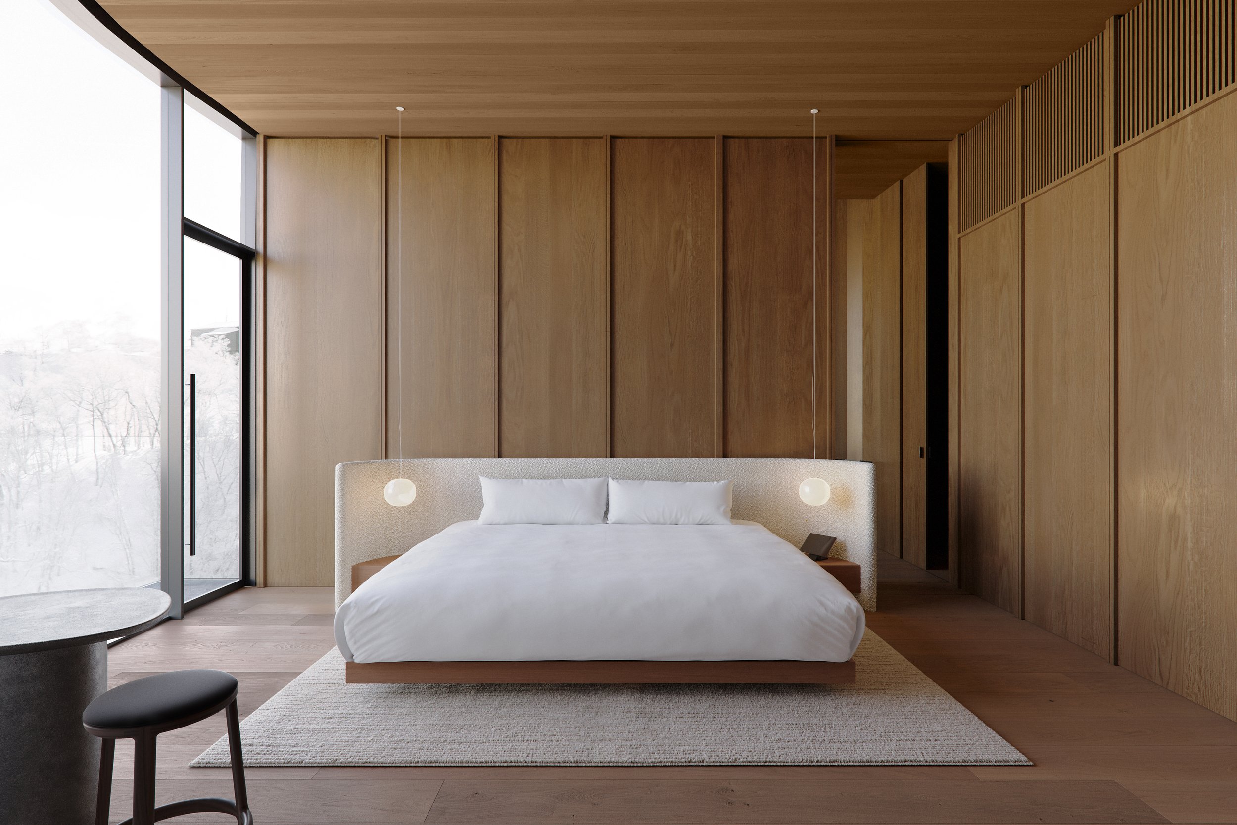 Modern bedroom with wooden walls and ceiling, large window, king-sized bed with white bedding and pillows, two hanging pendant lights, bedside table with lamp, and a small round table with stool near window.