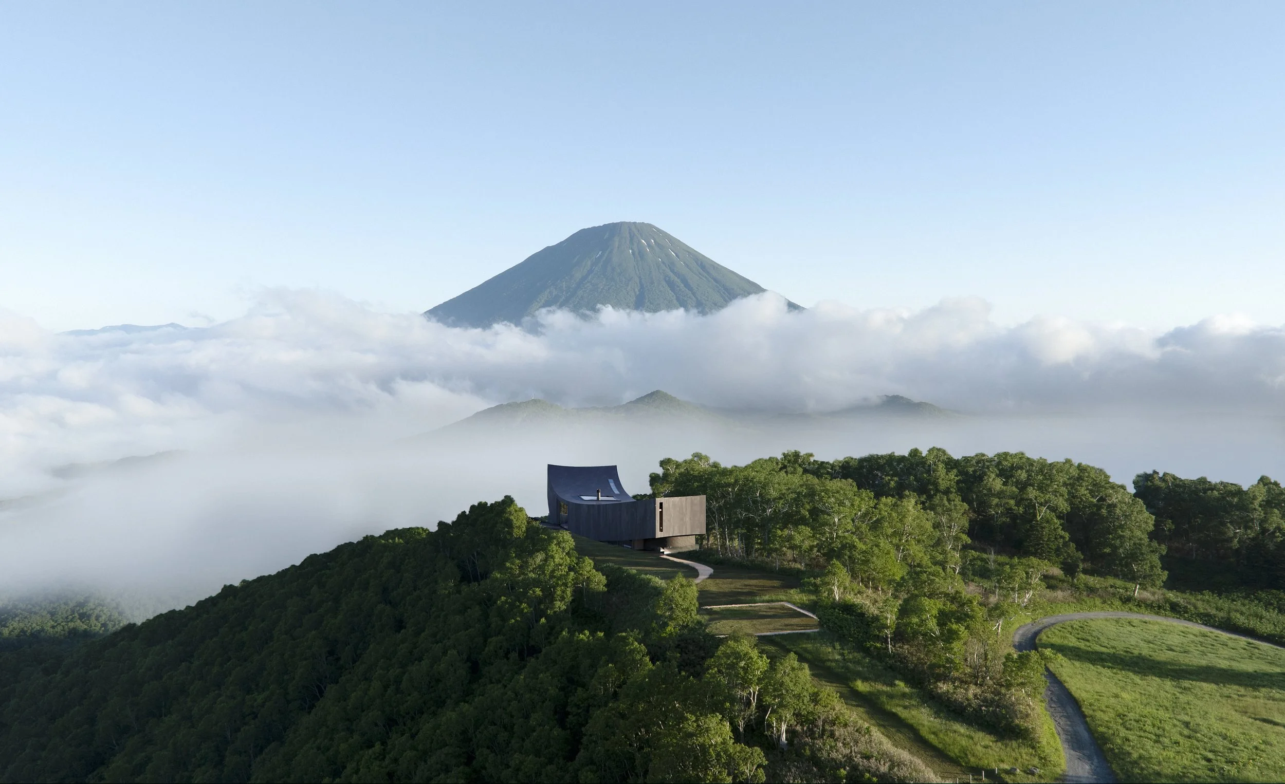 A modern building situated on a green hillside with dense trees, overlooking clouds, with a large volcanic mountain in the background.