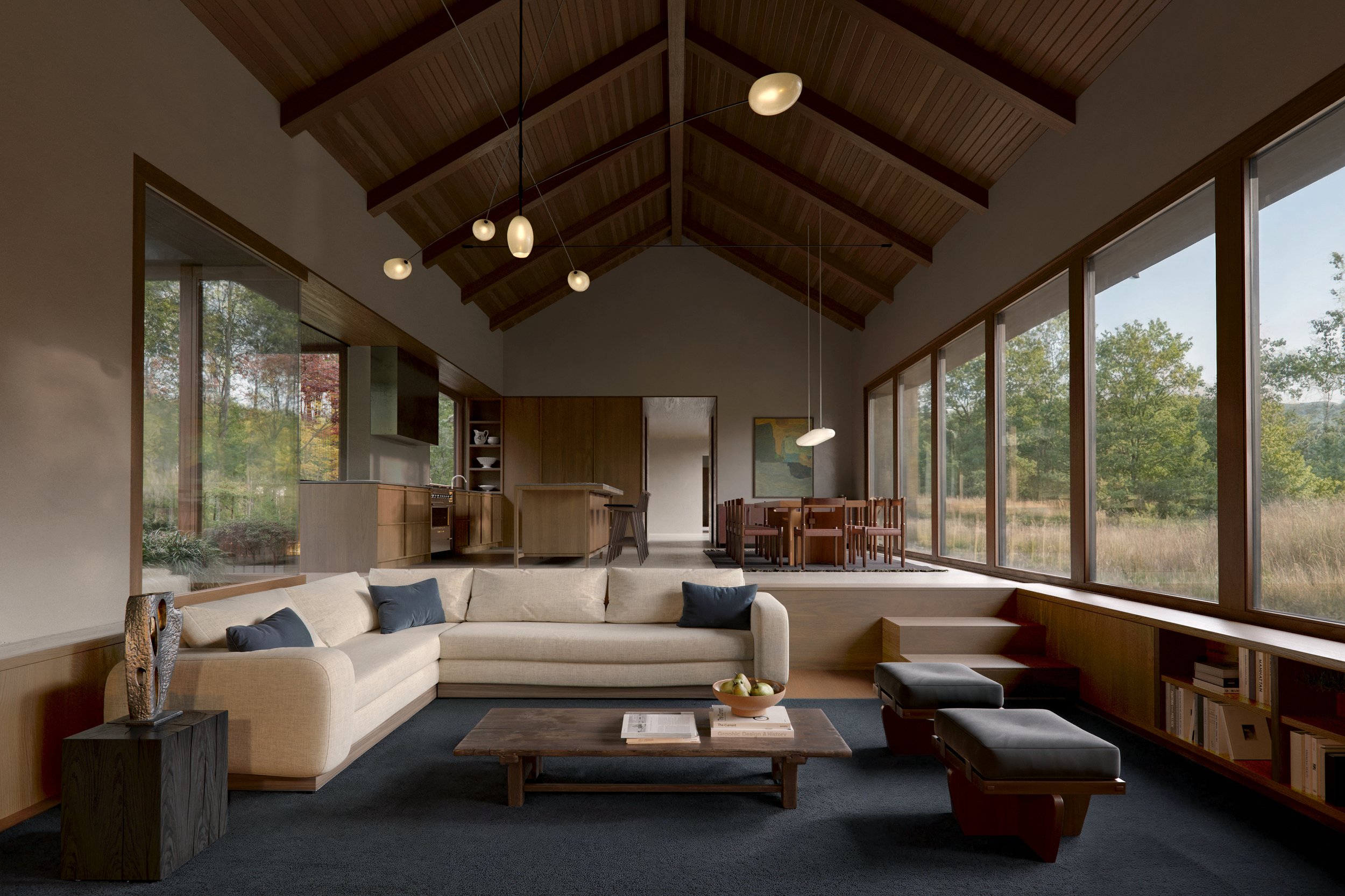 Living room with beige sofa, dark blue pillows, wooden coffee table with a fruit bowl, black ottoman, bookshelves under large windows showing trees outside, and a view of the dining area and kitchen in the background with wood accents and hanging light fixtures.