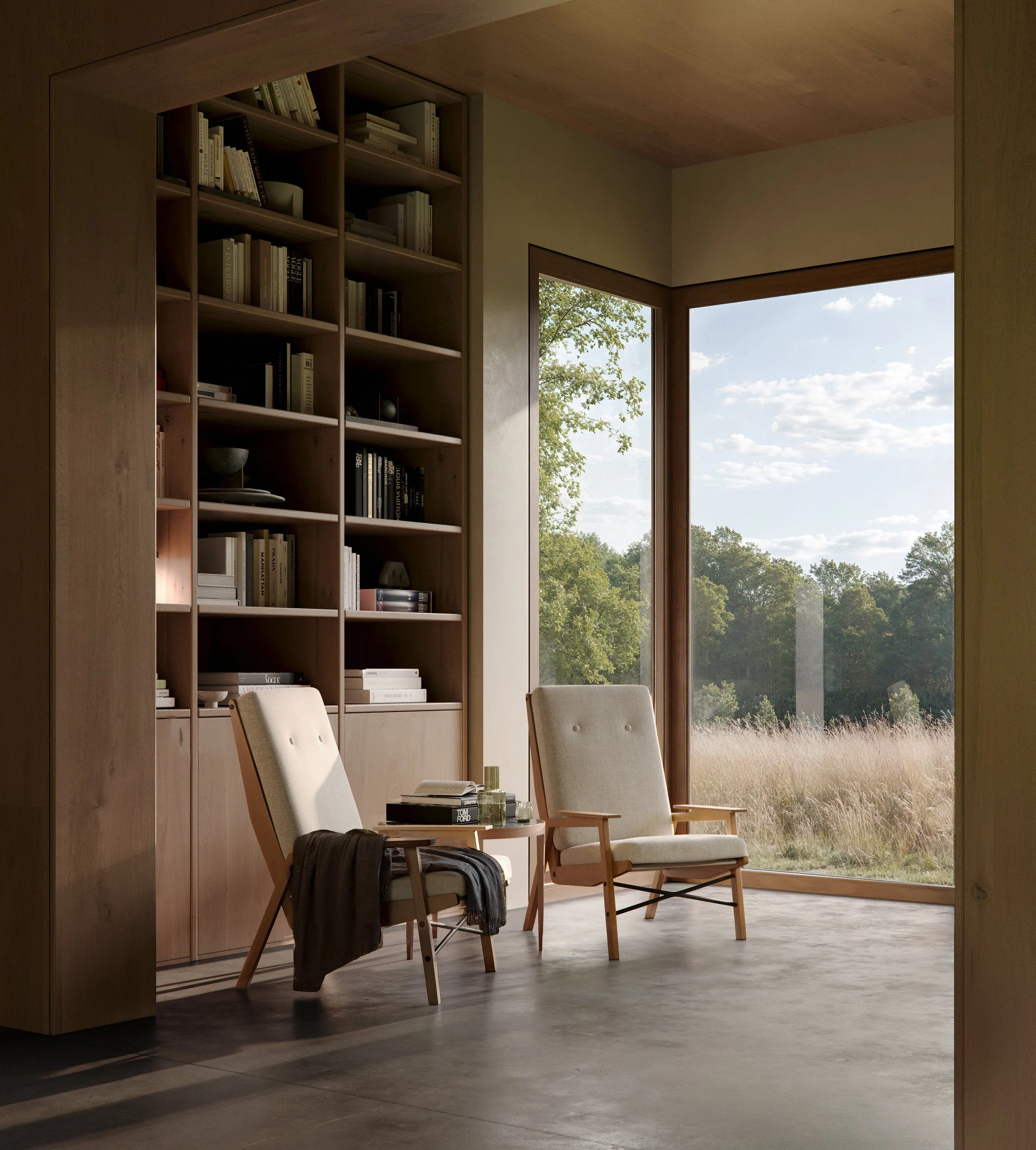 A cozy reading nook with two beige chairs and a small table in front, positioned by large windows overlooking a peaceful outdoor landscape with trees and grass, and a wall of bookshelves filled with books.