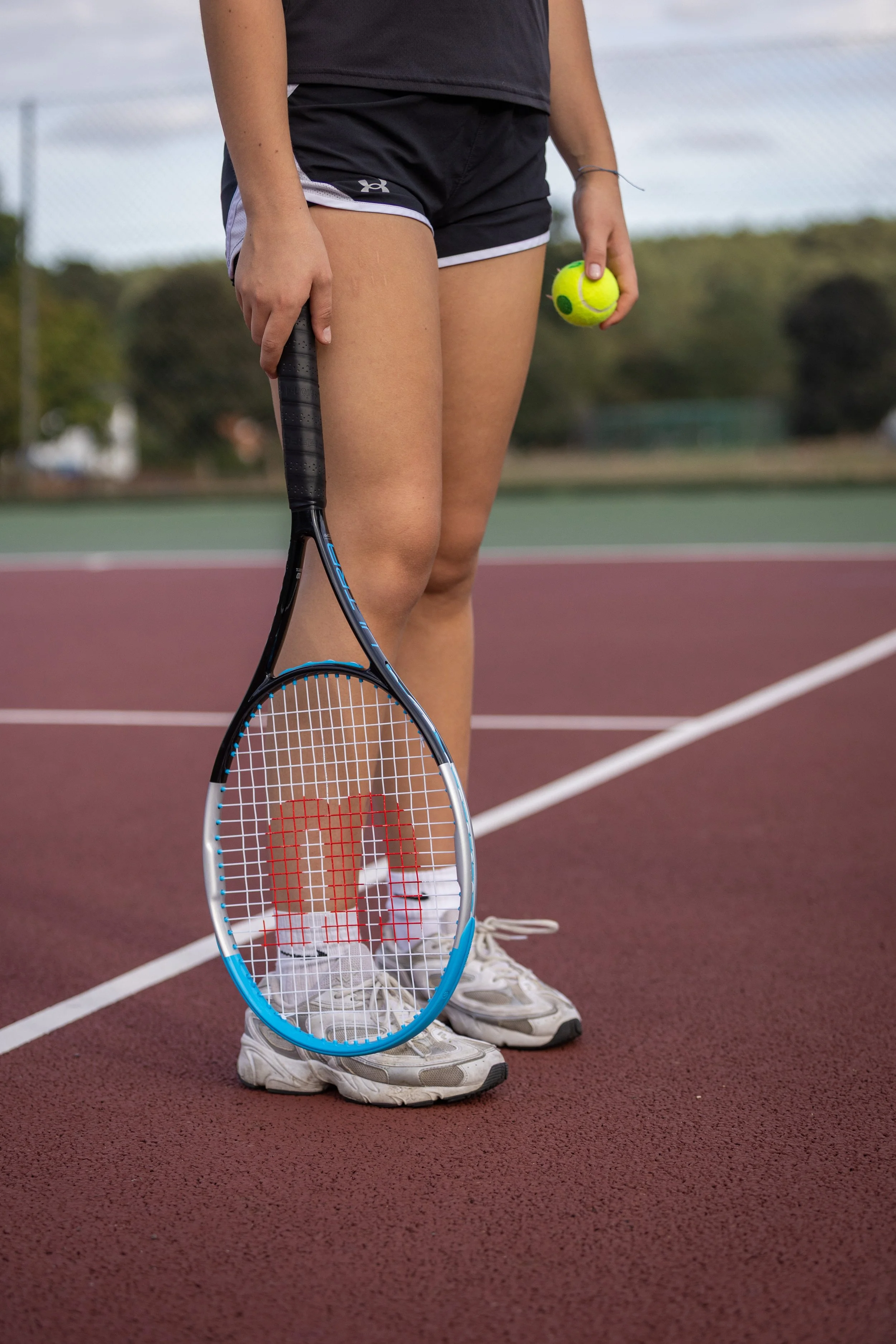 A person standing on a tennis court holding a tennis ball in their right hand and a tennis racket in their left, wearing athletic shoes, black shorts, and a black top.
