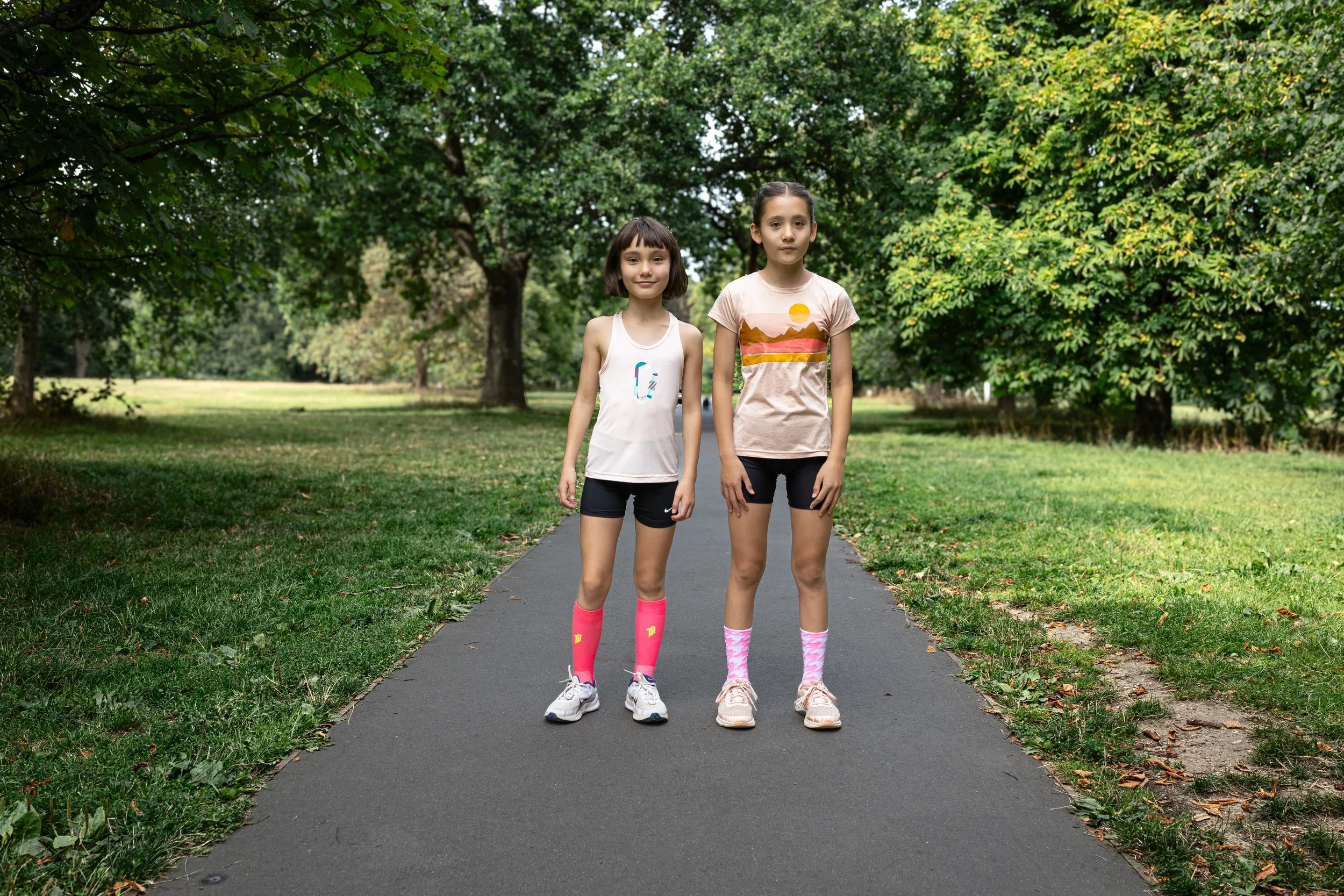 Two young girls standing on a paved walking trail in a park with trees and grass in the background.