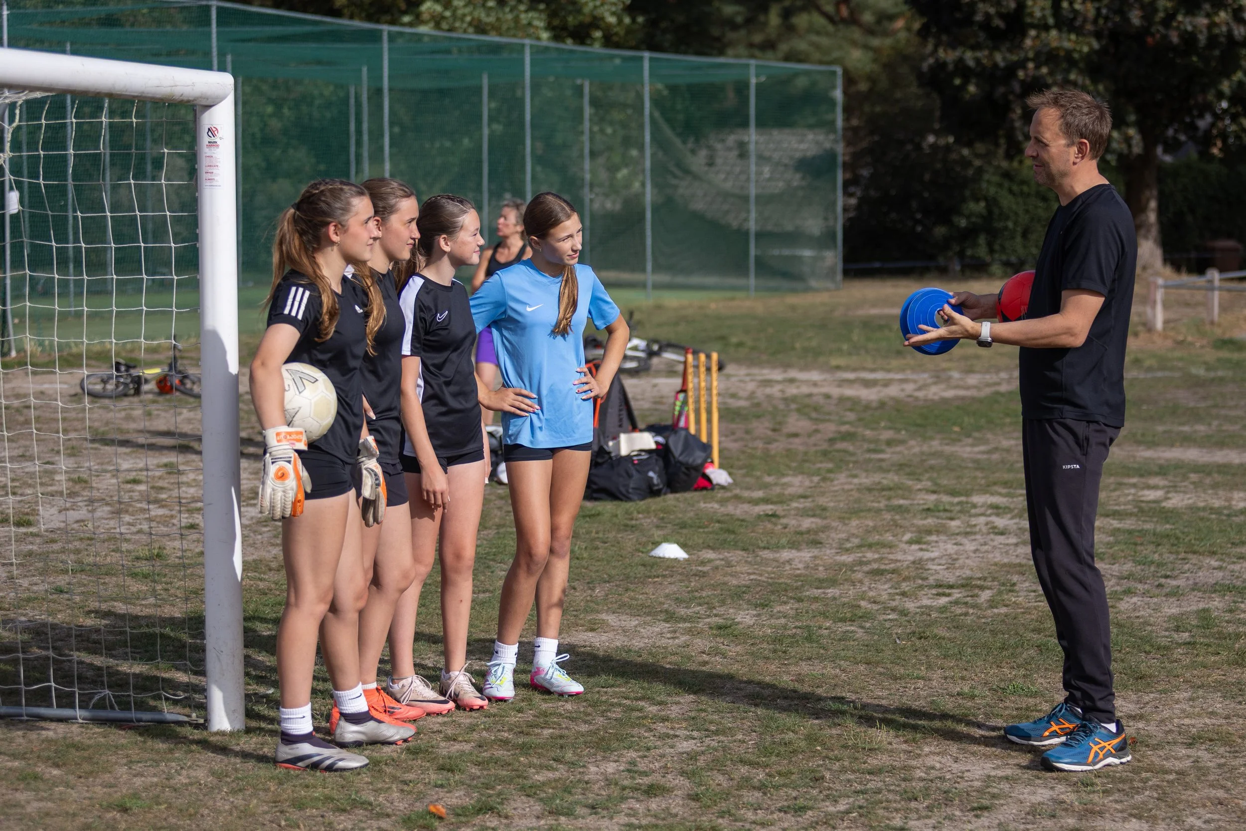 A coach giving instructions to a girls' soccer team during practice, outdoors on a soccer field.