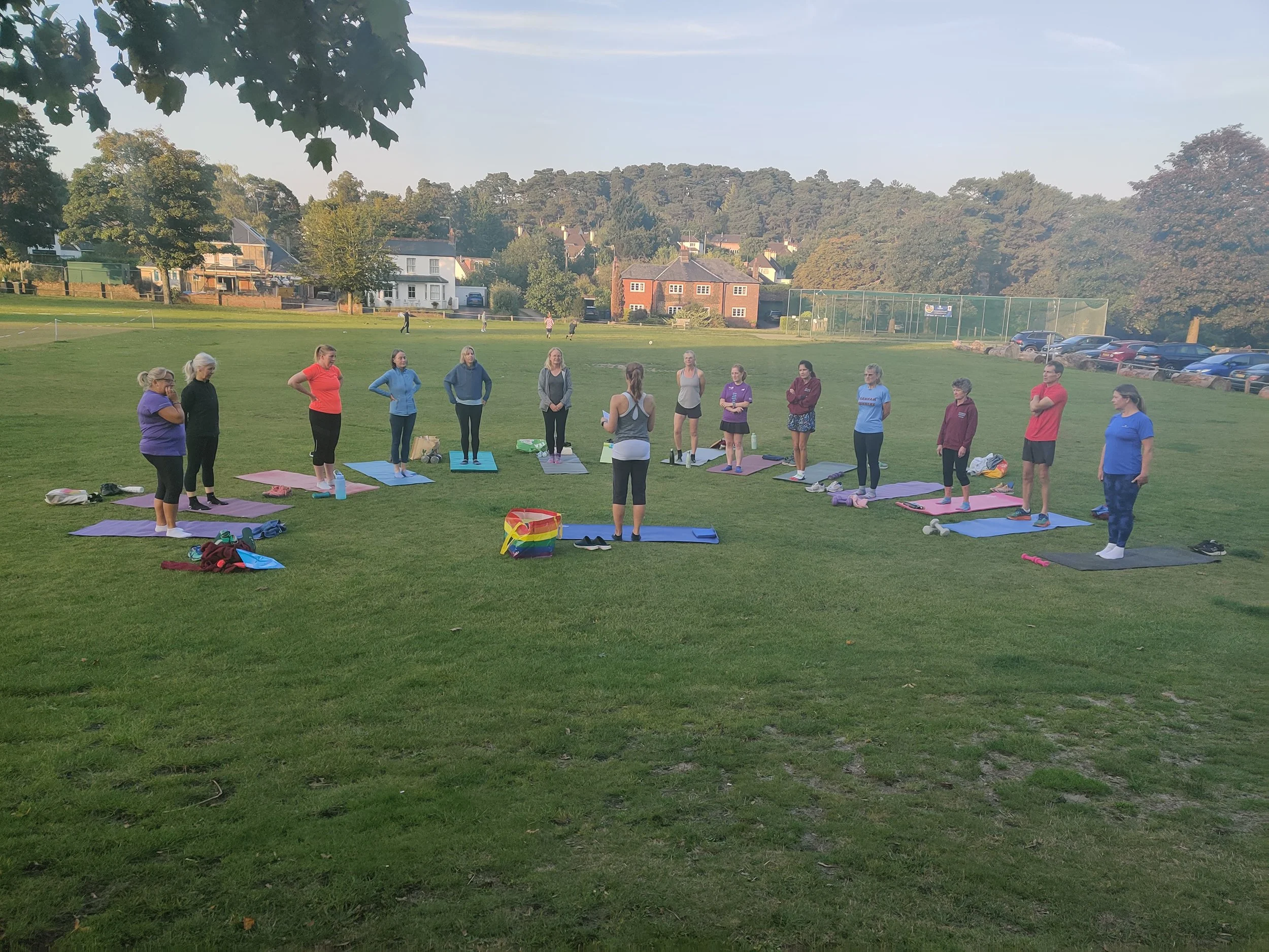 Group of people participating in an outdoor yoga class on a grassy field in the early morning or late afternoon, with trees and houses in the background.