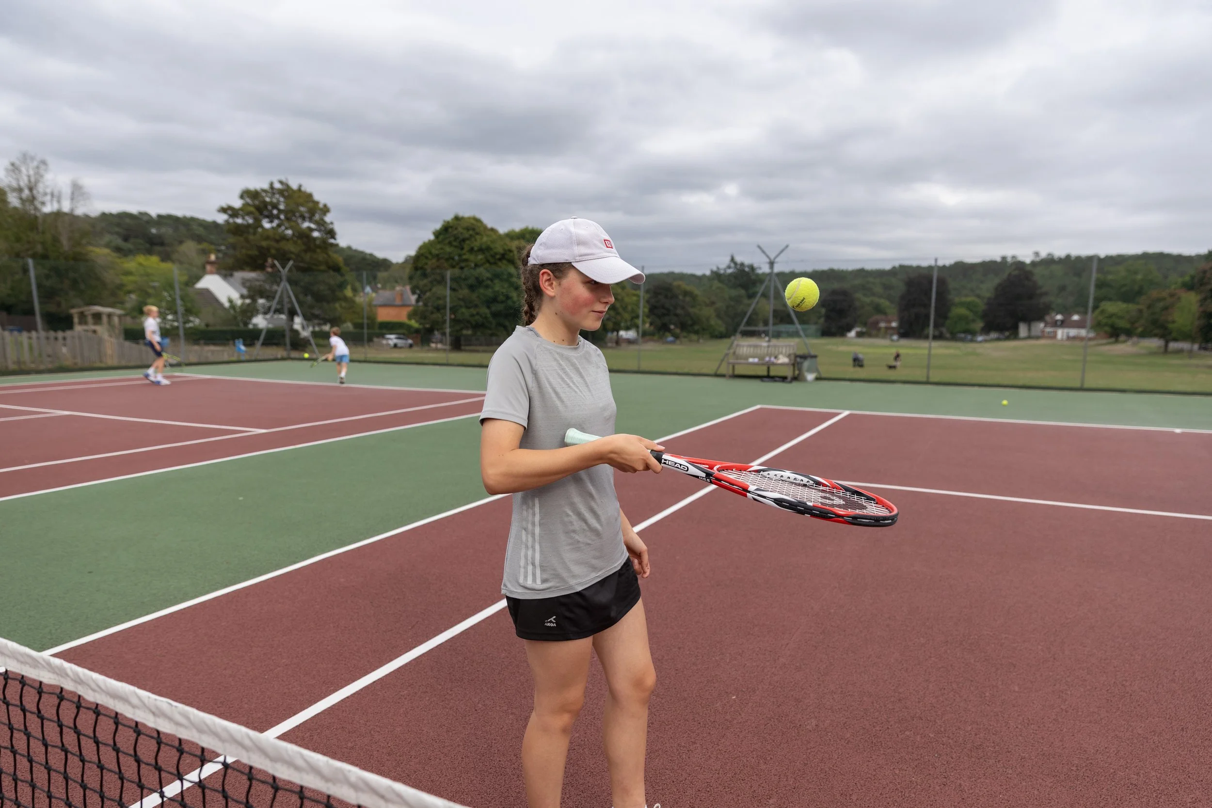 A young woman on a tennis court holding a tennis racket, with two children playing in the background and overcast skies above.