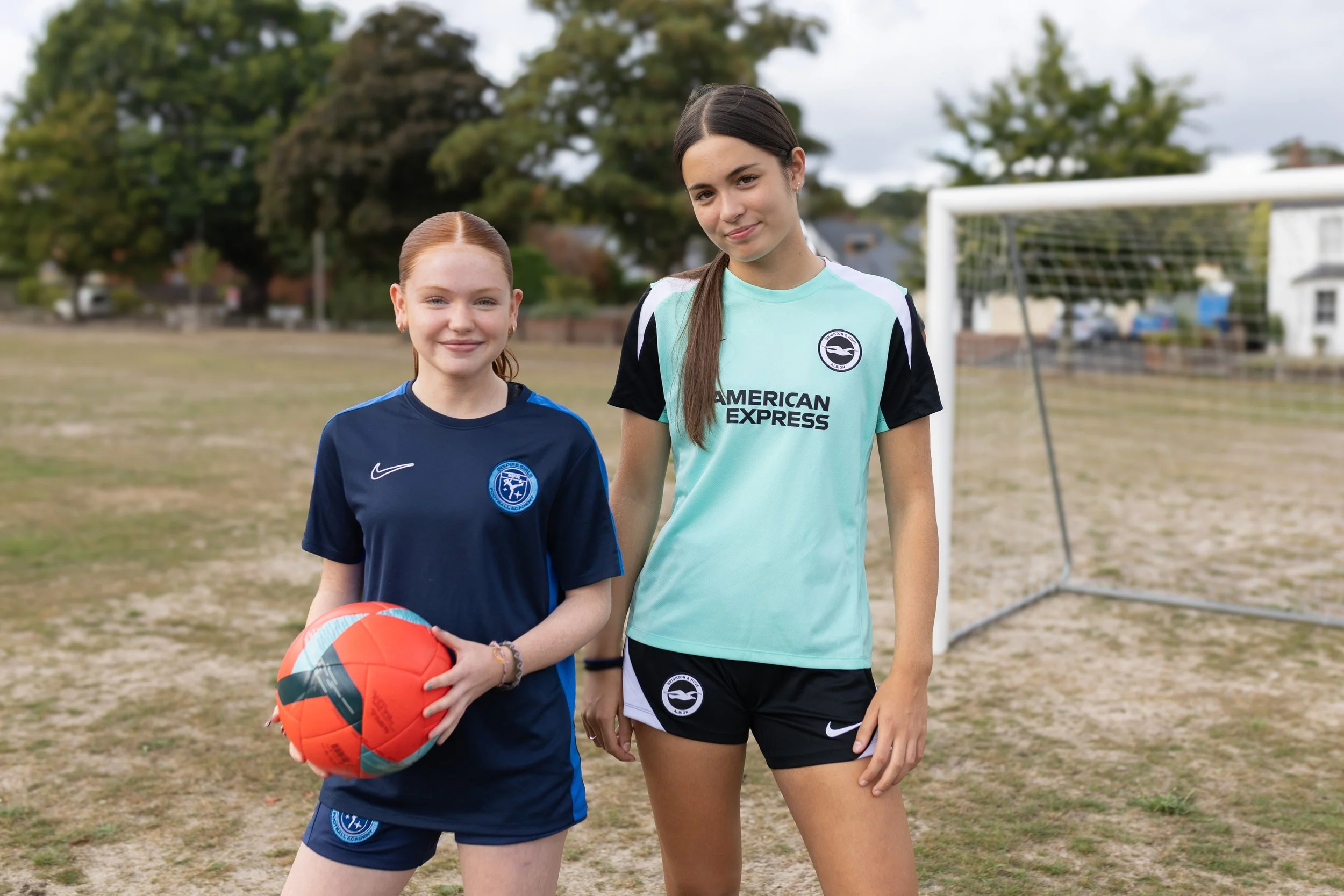 Two young women in soccer uniforms standing on a field, with a soccer goal in the background. One is holding a soccer ball.