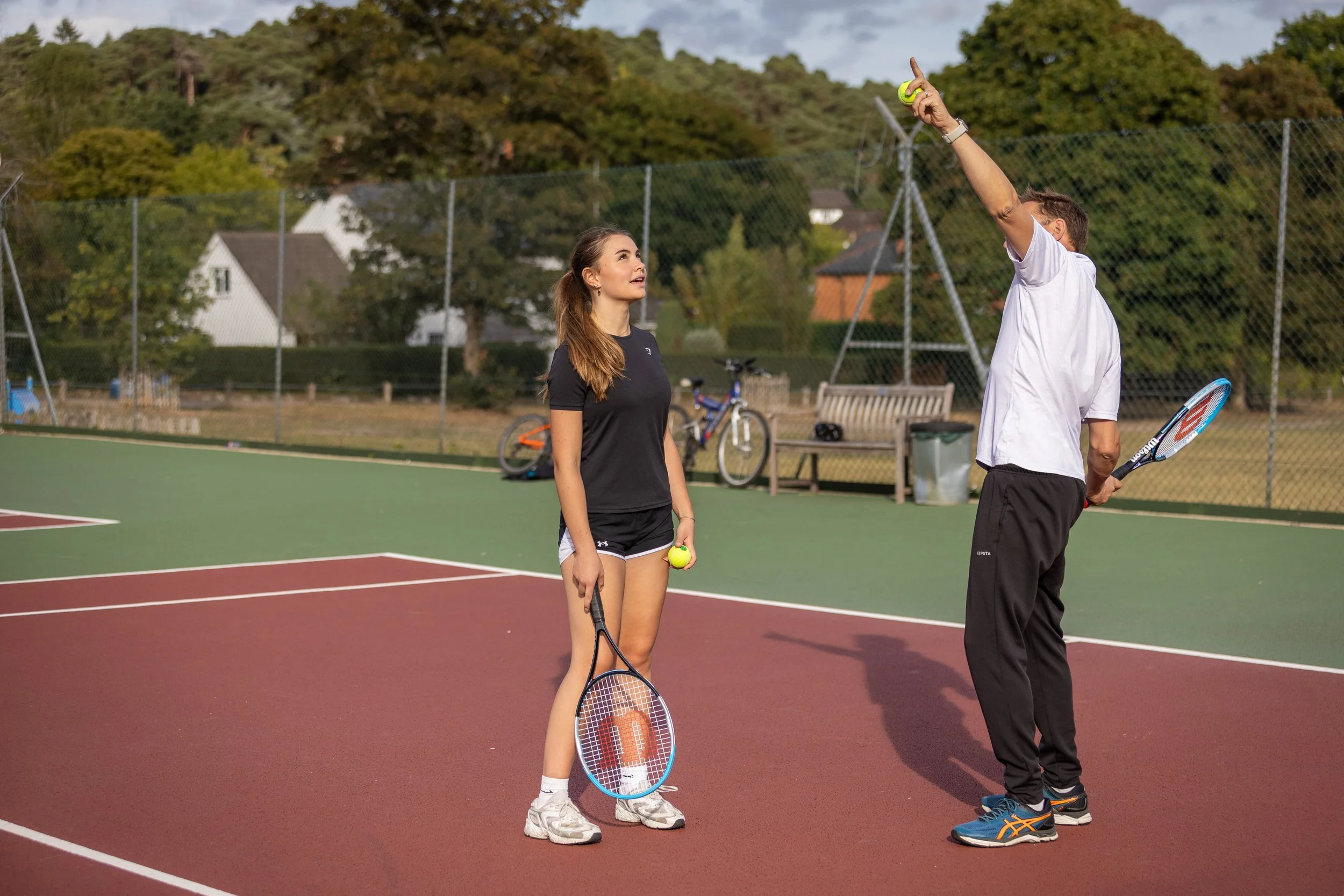A man and a woman on a tennis court, both holding tennis rackets, with the man pointing upwards and holding a tennis ball, and the woman looking at him, holding a tennis ball in her hand.