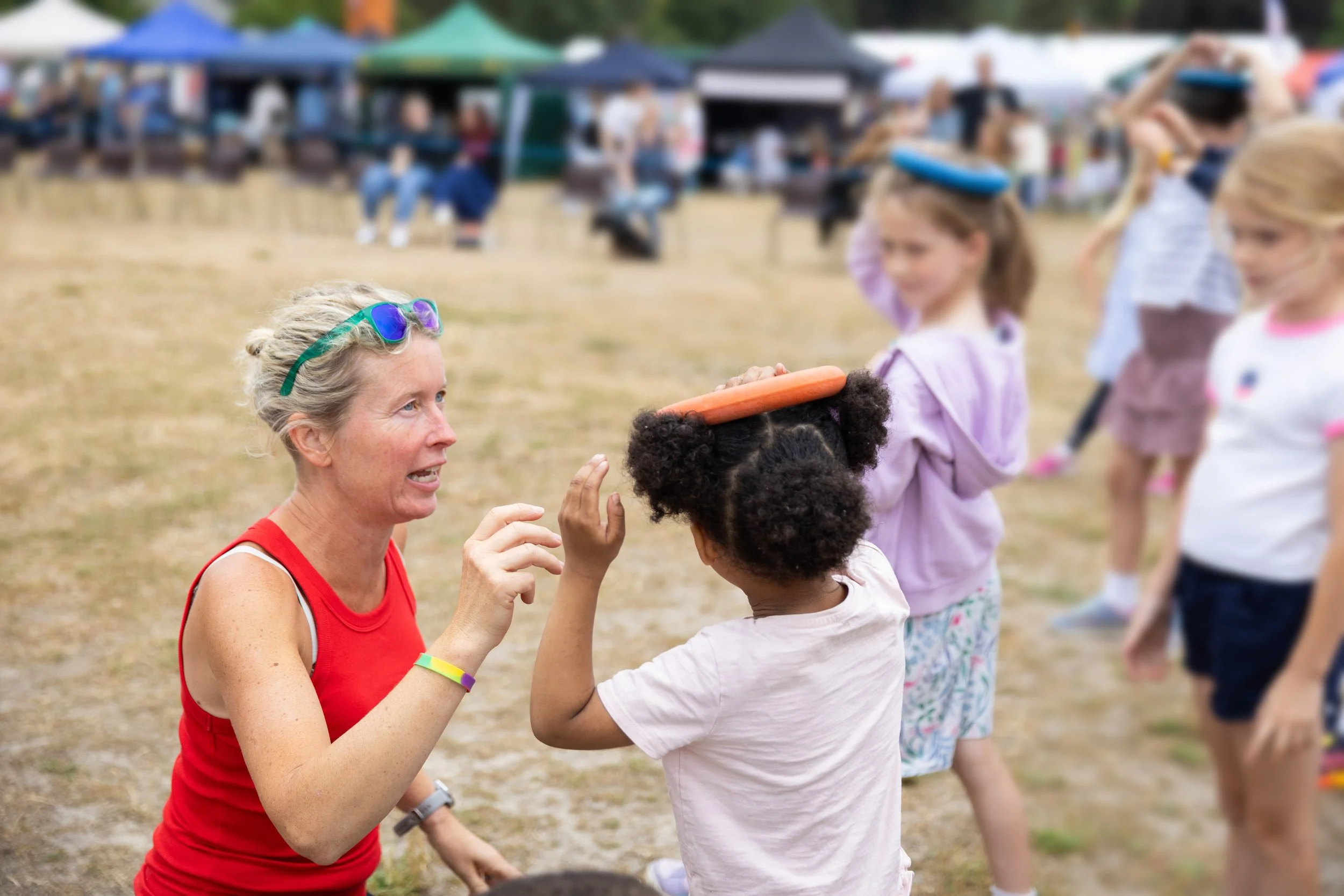 A woman in a red tank top talks to a young girl with an orange frisbee on her head at an outdoor event with children in line and tents in the background.