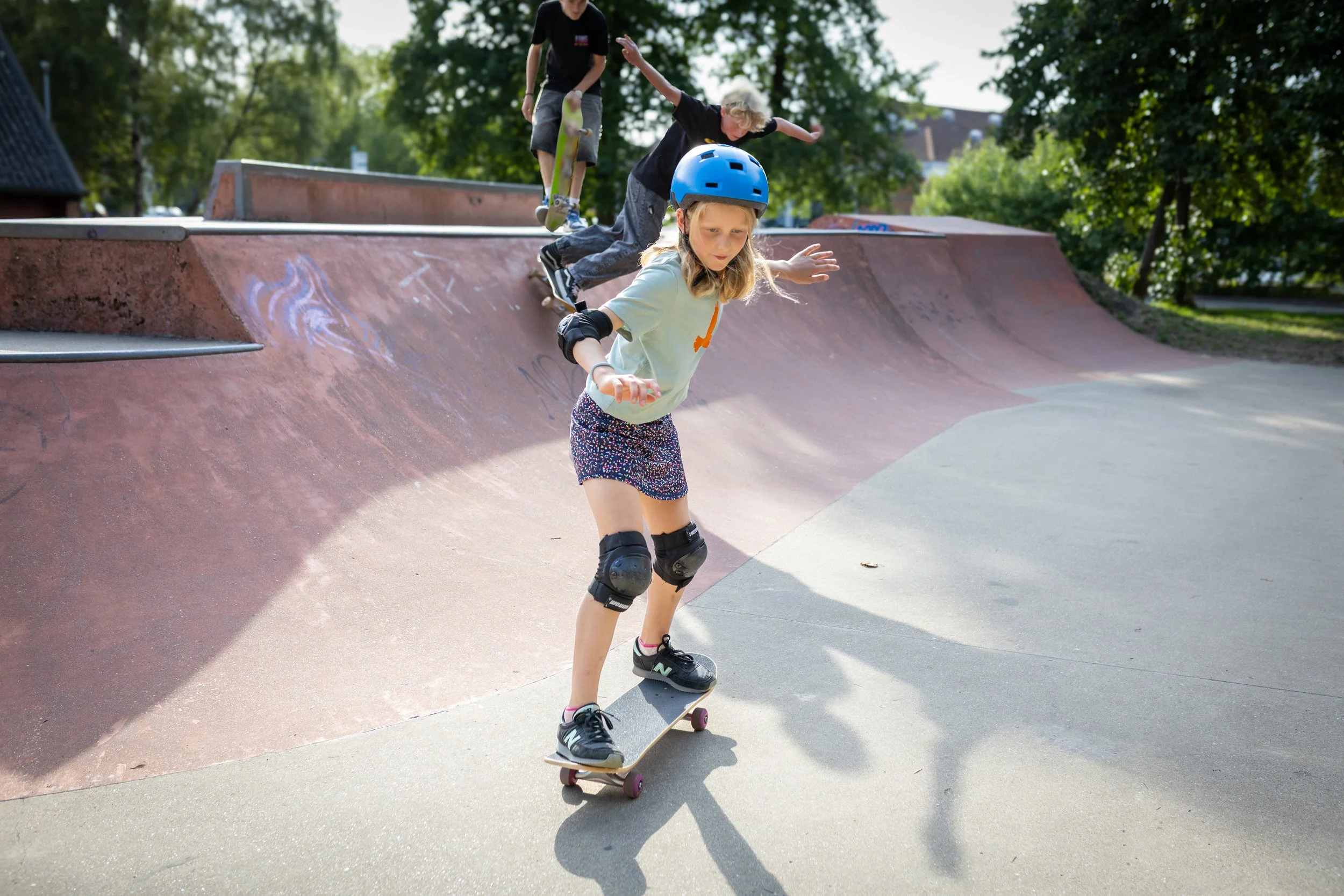 A young girl skateboarding at a skate park while wearing a blue helmet and knee pads, with two boys in the background also skateboarding.