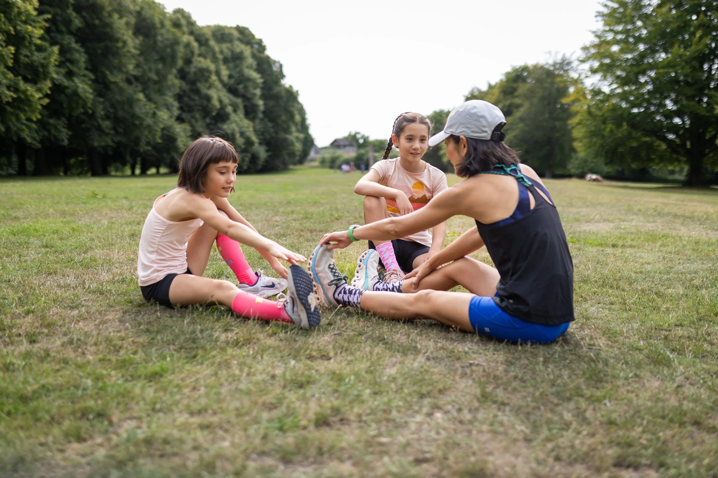 Three girls and an adult woman stretching on a grassy field in a park, sitting with legs extended and reaching toward their toes, surrounded by trees.