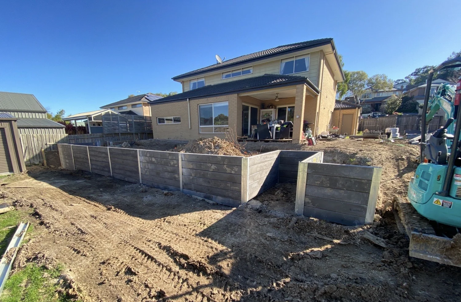 Concrete sleeper retaining wall being built with an excavator behind a new home