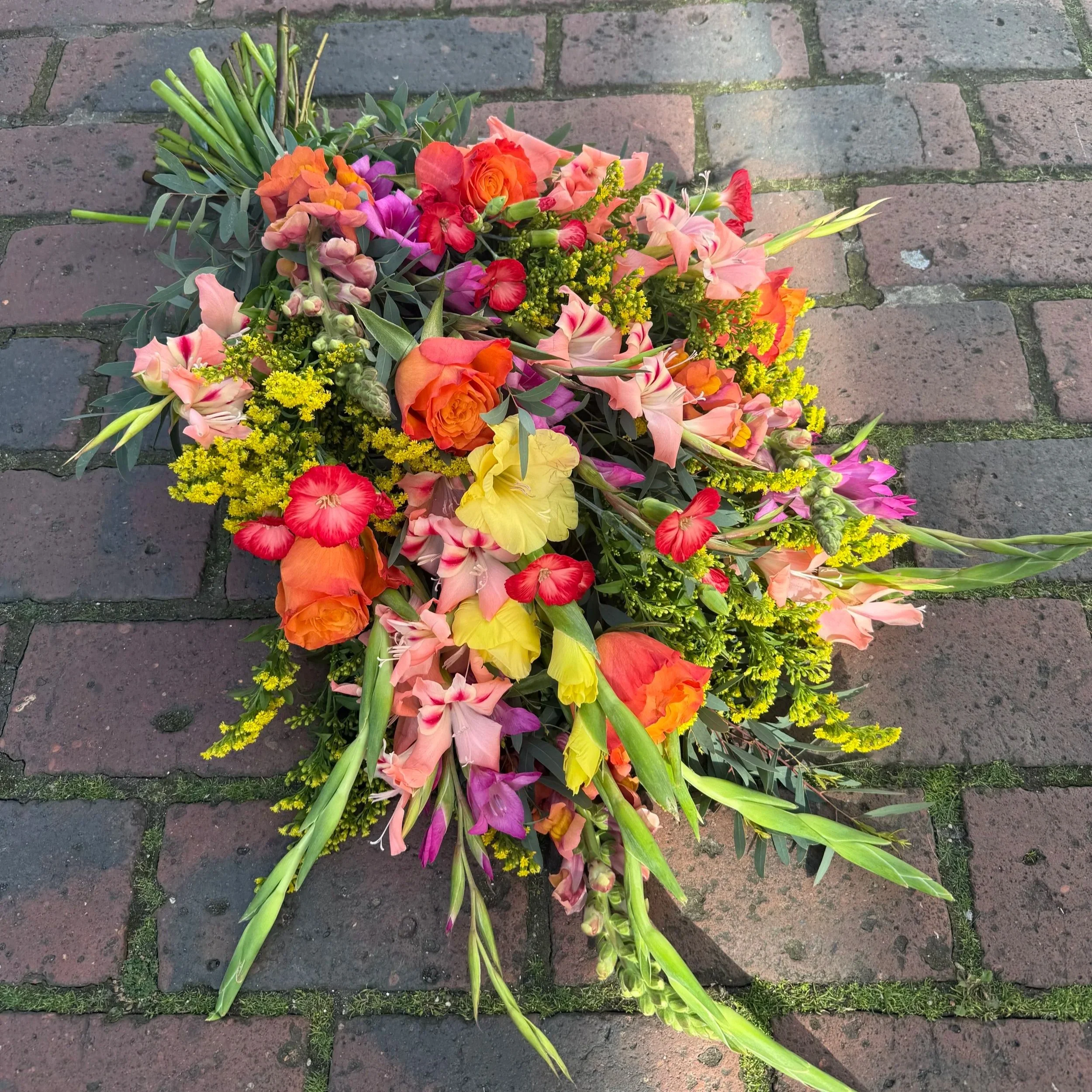 A colourful tied sheaf with various pink, orange, yellow, red, and pinks flowers along with green foliage, laid on a brick pathway.