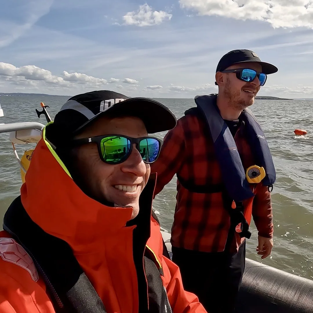 Two men on a boat enjoying a day on the water. They are wearing sunglasses, life jackets, and outdoor gear, with one smiling at the camera and the other standing near the water with a smile. The ocean and a partly cloudy sky are visible in the background.