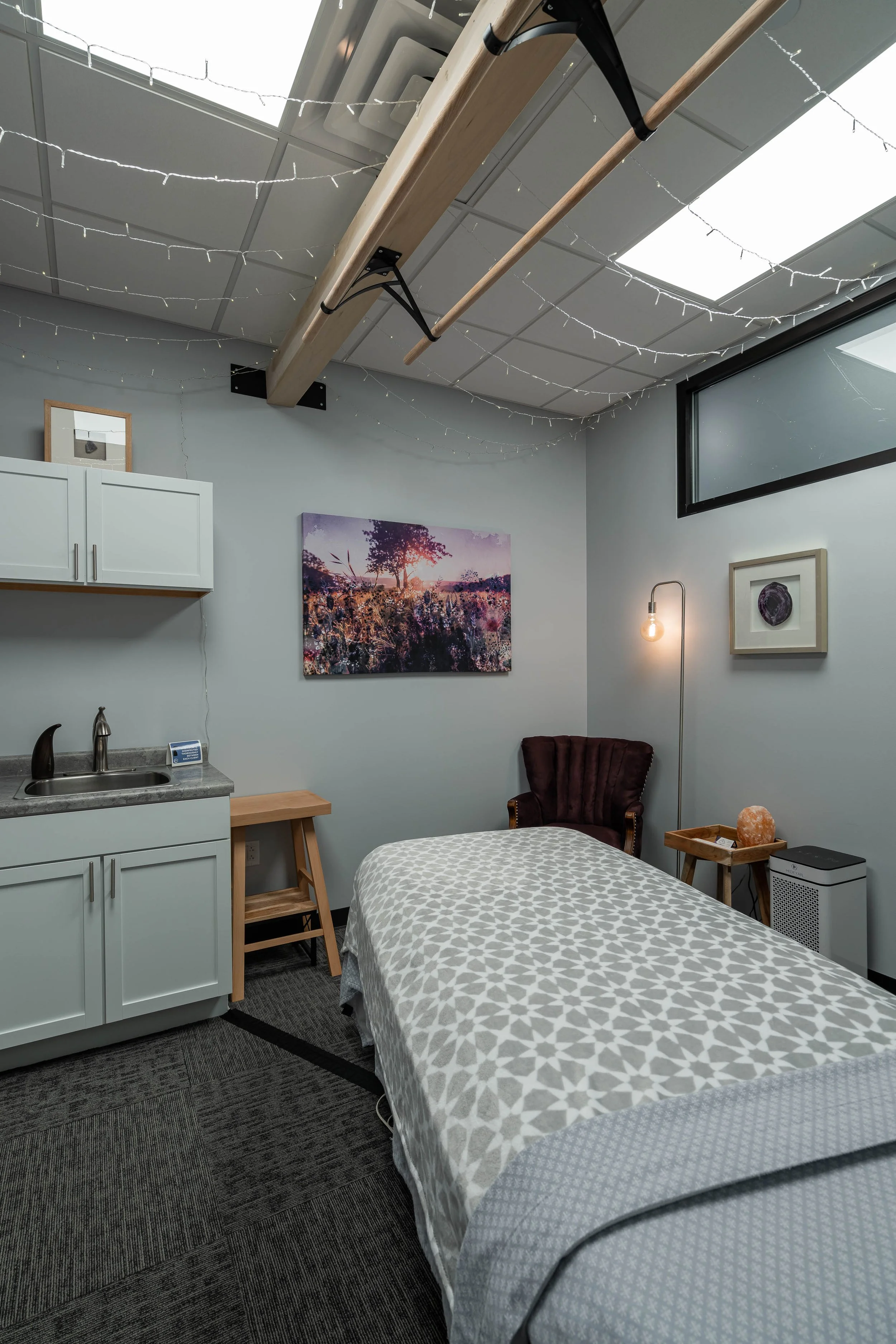 A cozy room with a bed covered in a patterned blanket, a brown armchair, a small wooden side table, a floor lamp with an exposed bulb, wall artworks including a landscape and a purple decoration, a white cabinet with a small framed picture, a sink ar